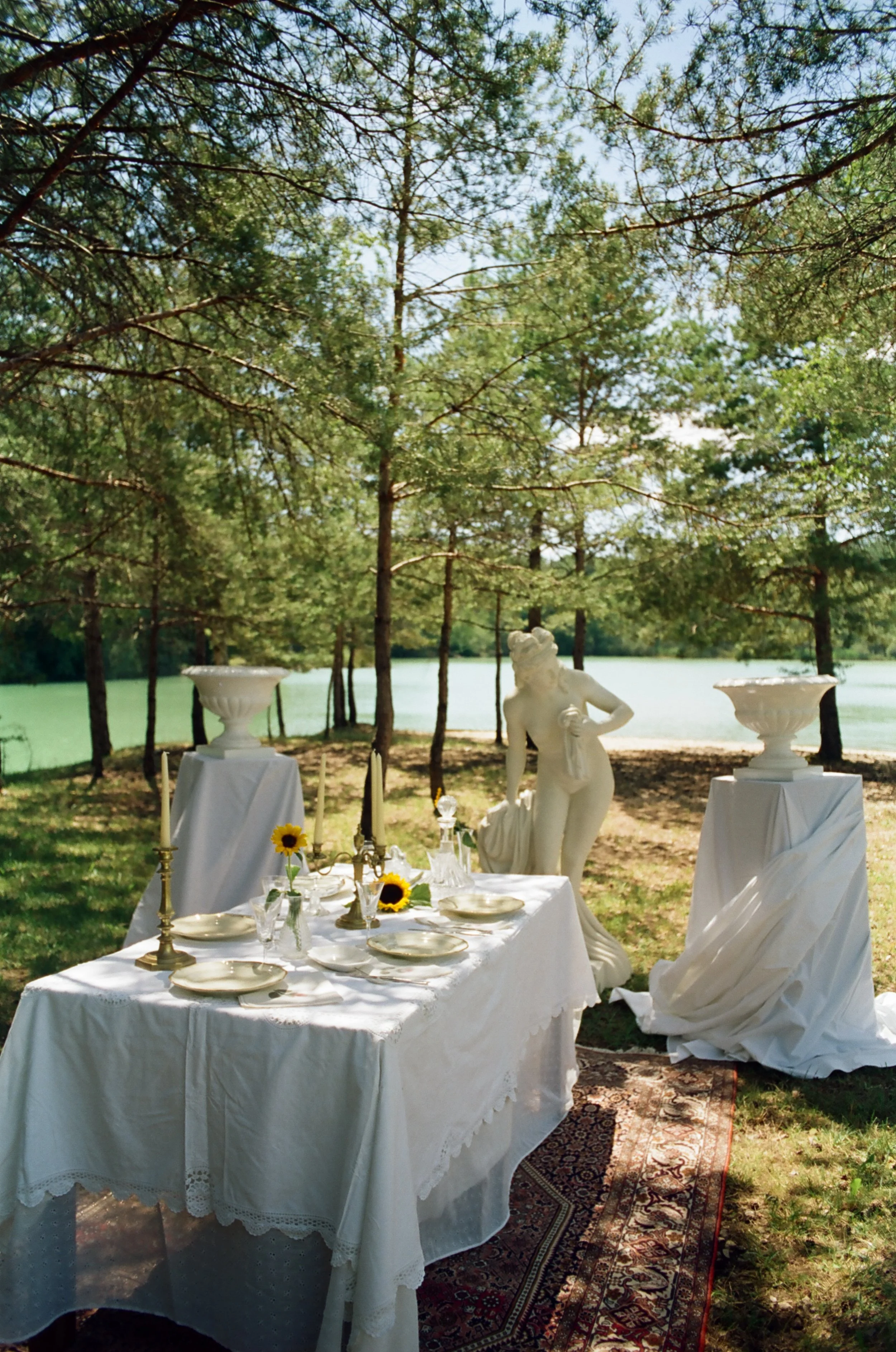 Table de style élégante décorée avec une nappe blanche, vaisselle, chandeliers et tournesols, située en plein air sous des arbres près d'un lac, avec une sculpture de femme en marbre dans le décor.