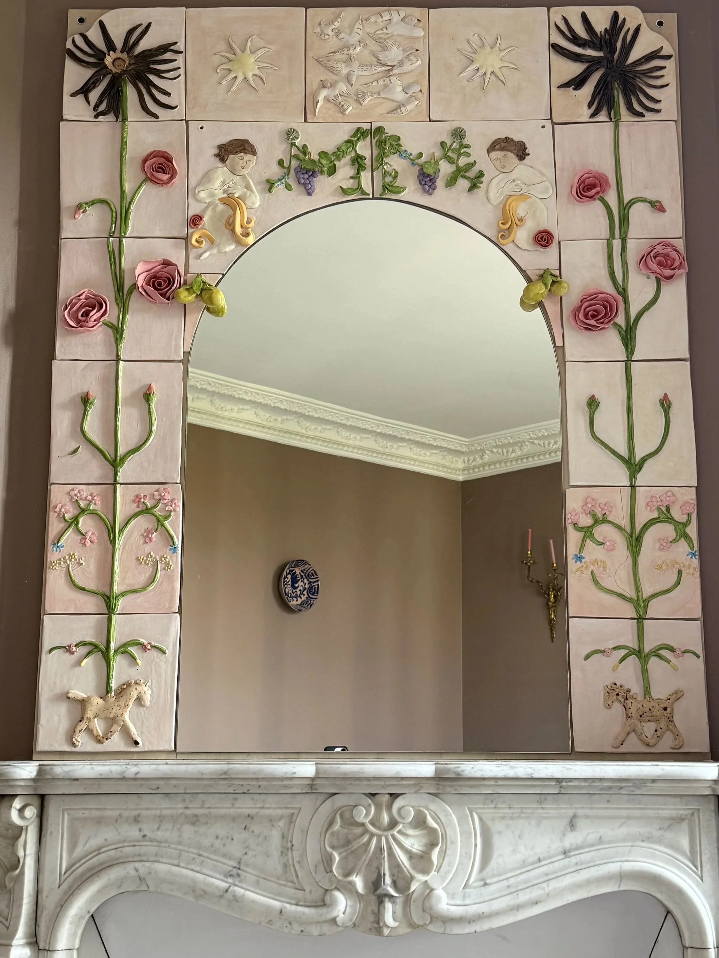 Miroir encadré de carreaux décoratifs illustrant des thèmes floraux et animaliers, avec des motifs de fleurs, de vignes, de personnages, d'oiseaux, de branchages, et d'animaux comme un cheval et un léopard en relief.