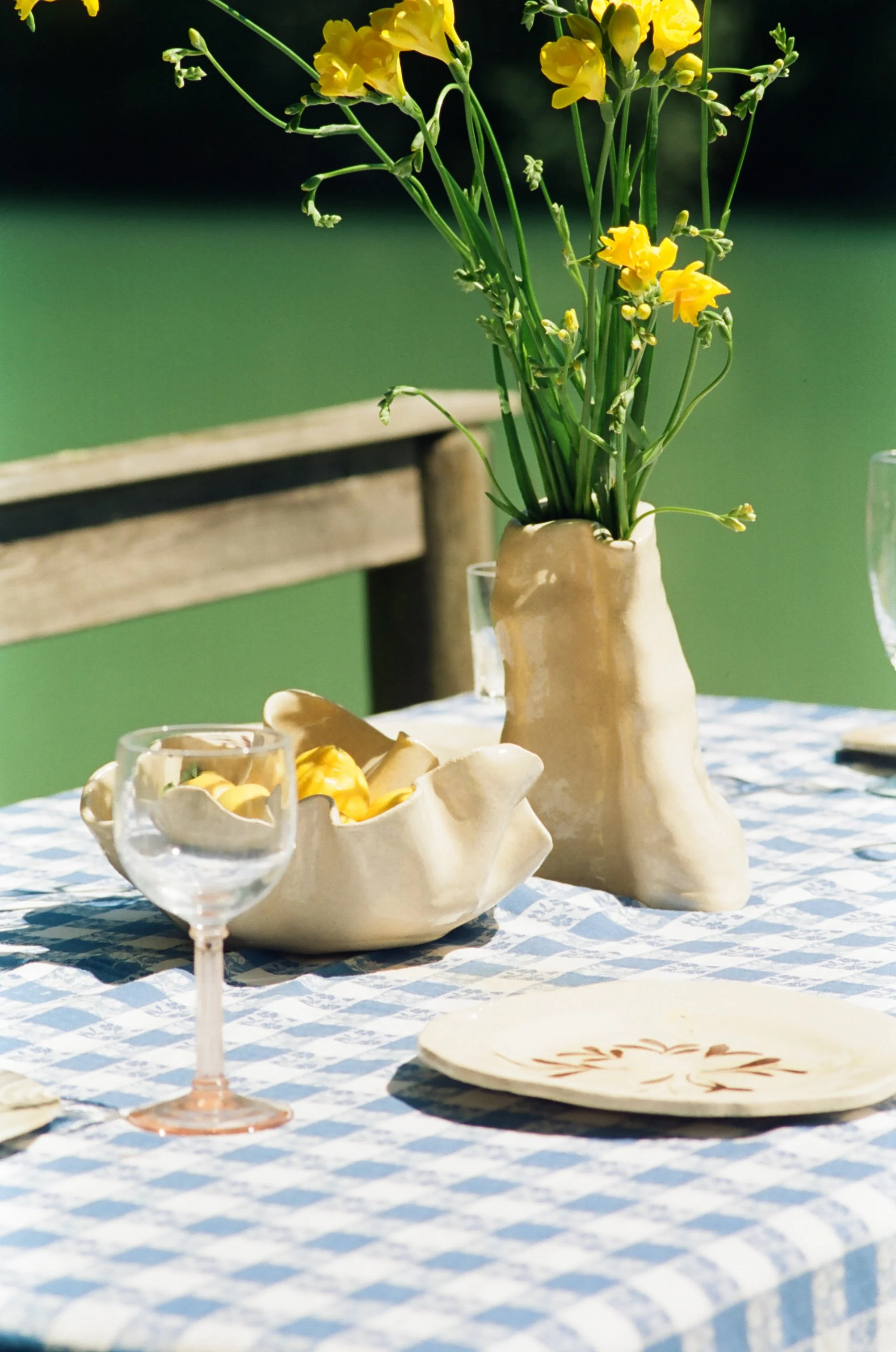 Une table dressée avec une nappe bleue et blanche à carreaux, décorée d'un vase en céramique contenant des fleurs jaunes, d'une coupelle en céramique avec des jaunes en chocolat, une assiette ornementée en céramique, et des verres à vin vides, en plein air avec un fond de verdure.
