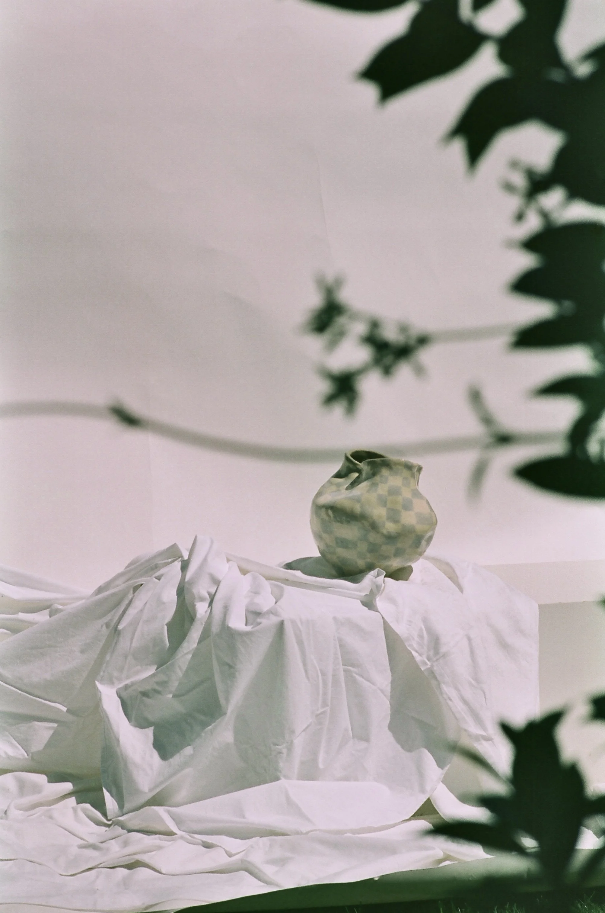 Une vase en céramique verte avec un motif en damier clair, placée sur un tissu blanc froissé, avec des branches de feuillage floues en premier plan.