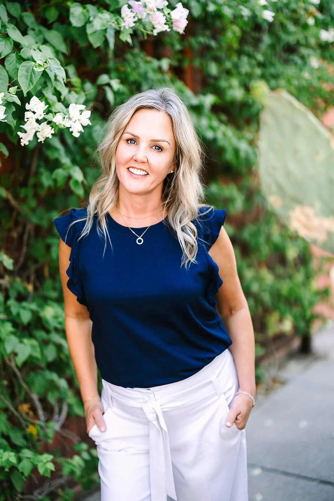 Jen with blonde hair, wearing a navy top and white pants, standing outdoors with greenery and white flowers in the background.