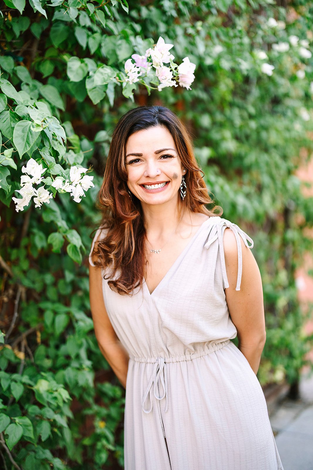 Jessica with long wavy brown hair and a bright smile standing outdoors in front of lush green foliage with white and pink flowers, wearing a light-colored sleeveless dress with tied shoulder straps.