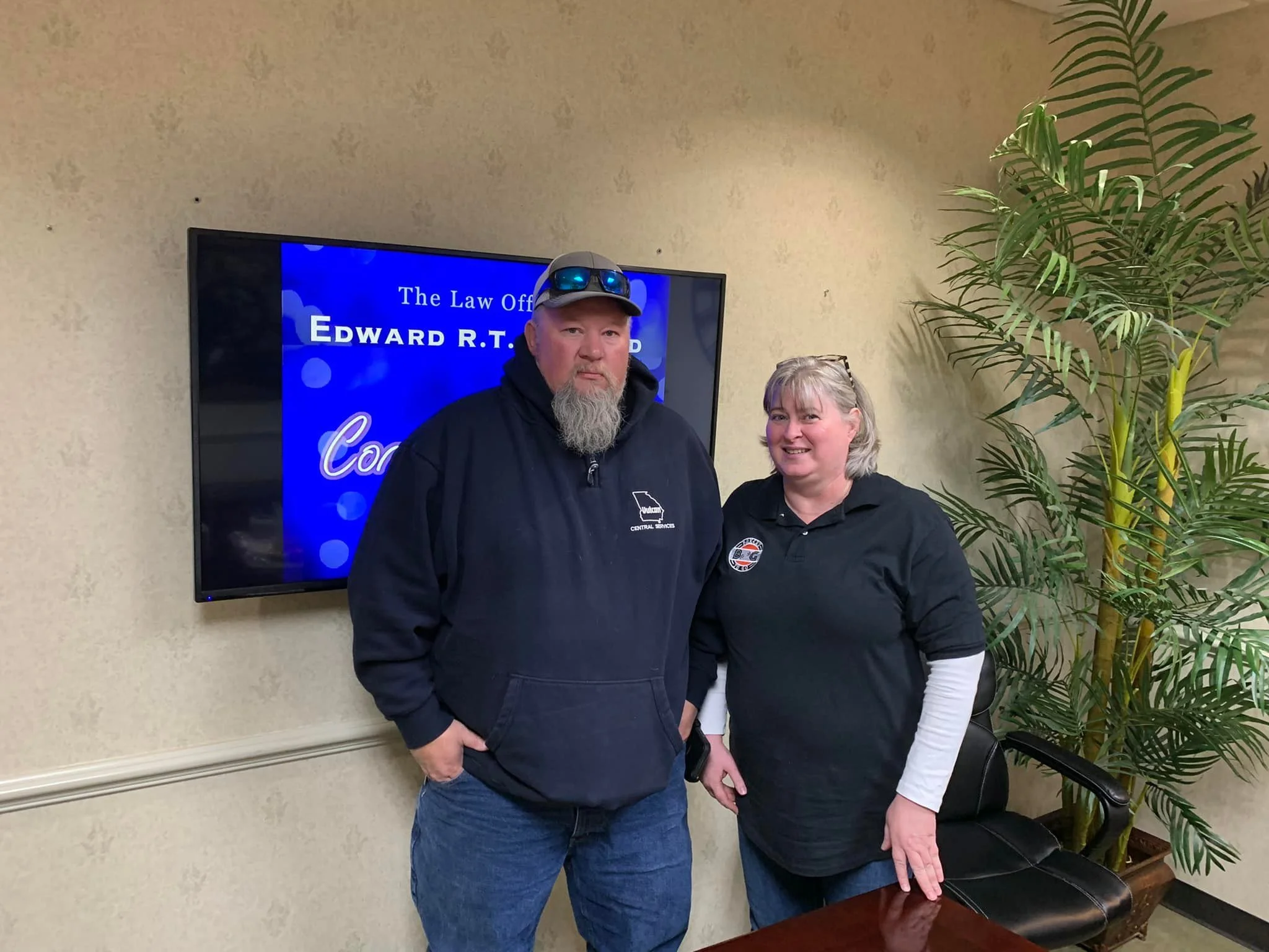 A man and woman standing side by side indoors, with a large flat-screen TV behind them displaying a blue screen with text about a law office. The man has a beard, is wearing a black hoodie and sunglasses on his head, and has his hands in his pockets.