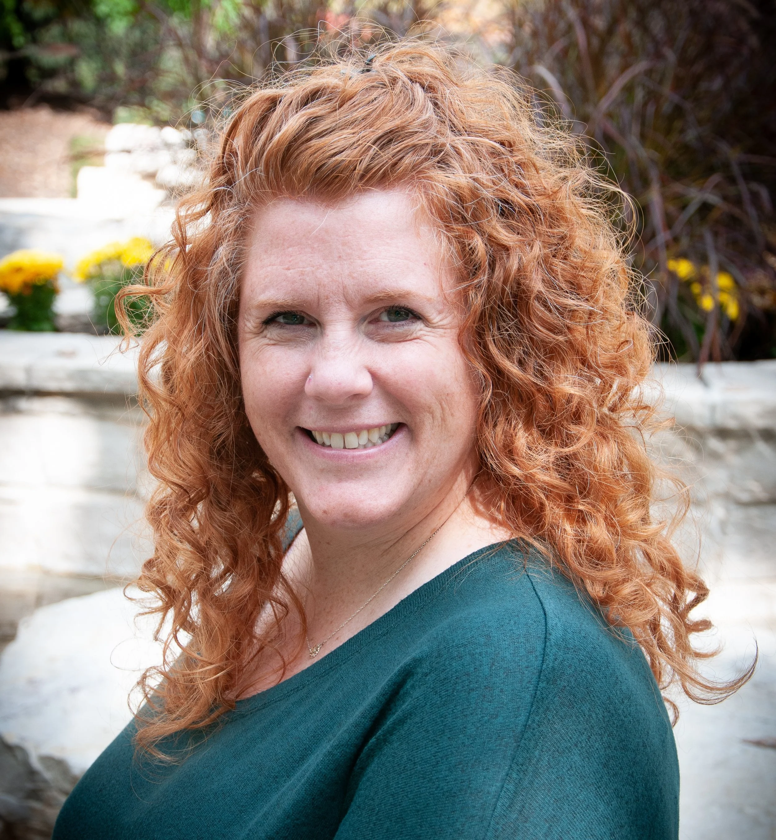 A woman with curly red hair smiling outdoors in front of a stone wall and plants.