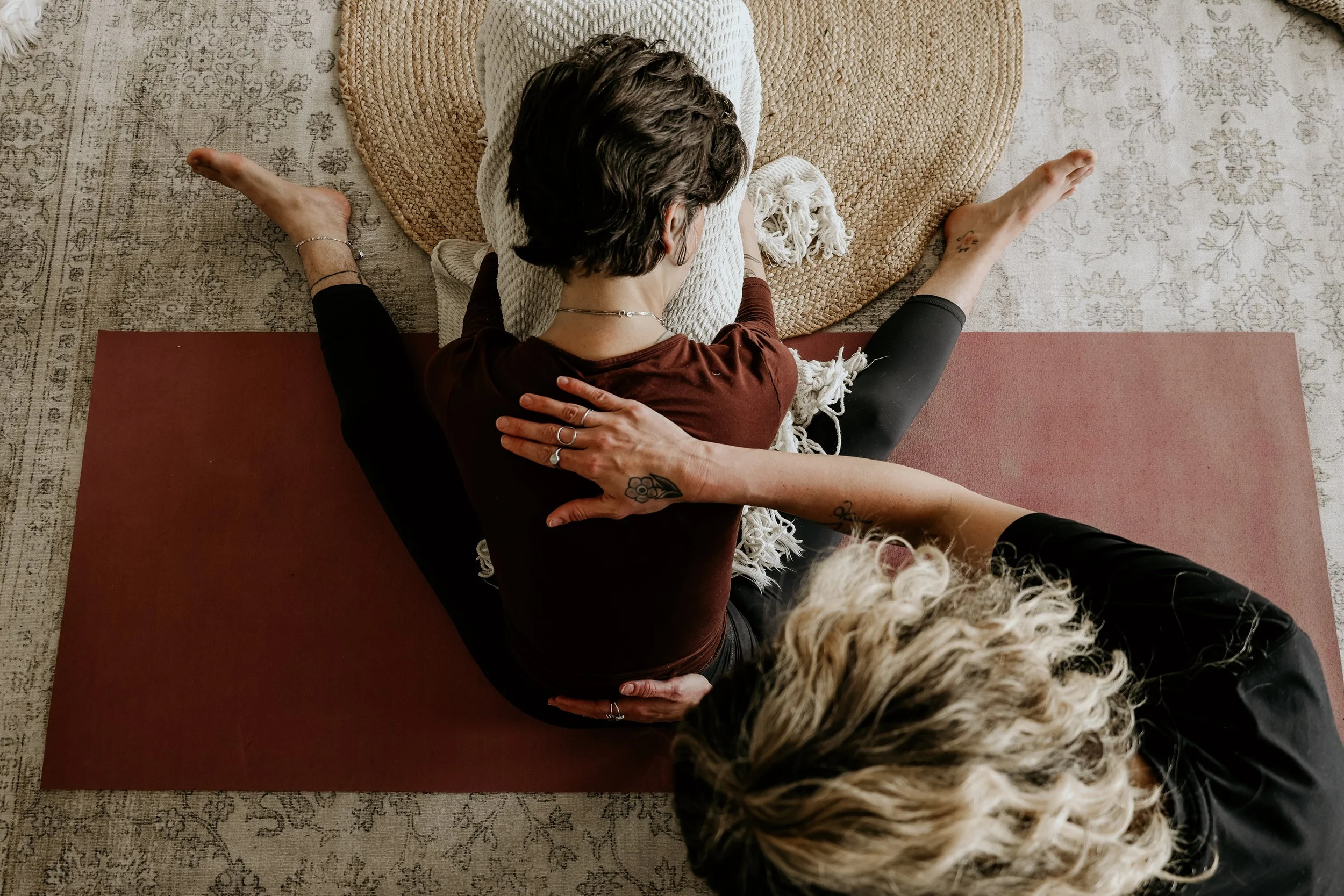 Indoor yoga therapy session, client in a supported forward fold on a red yoga mat and the other supporting from behind, with a beige rug and woven round mat underneath.
