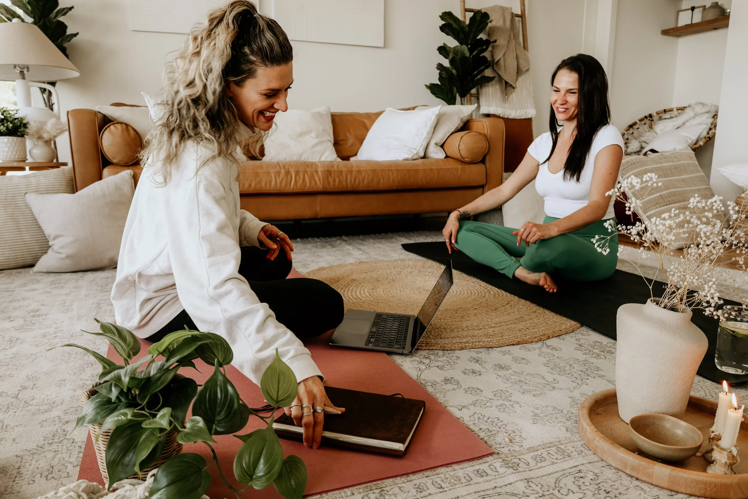 Two women sitting on the floor of a cozy living room, engaging in a relaxed, cheerful conversation. One woman with curly hair touches a book, while the other with straight hair sits on a yoga mat near a laptop, all surrounded by comfortable pillows,