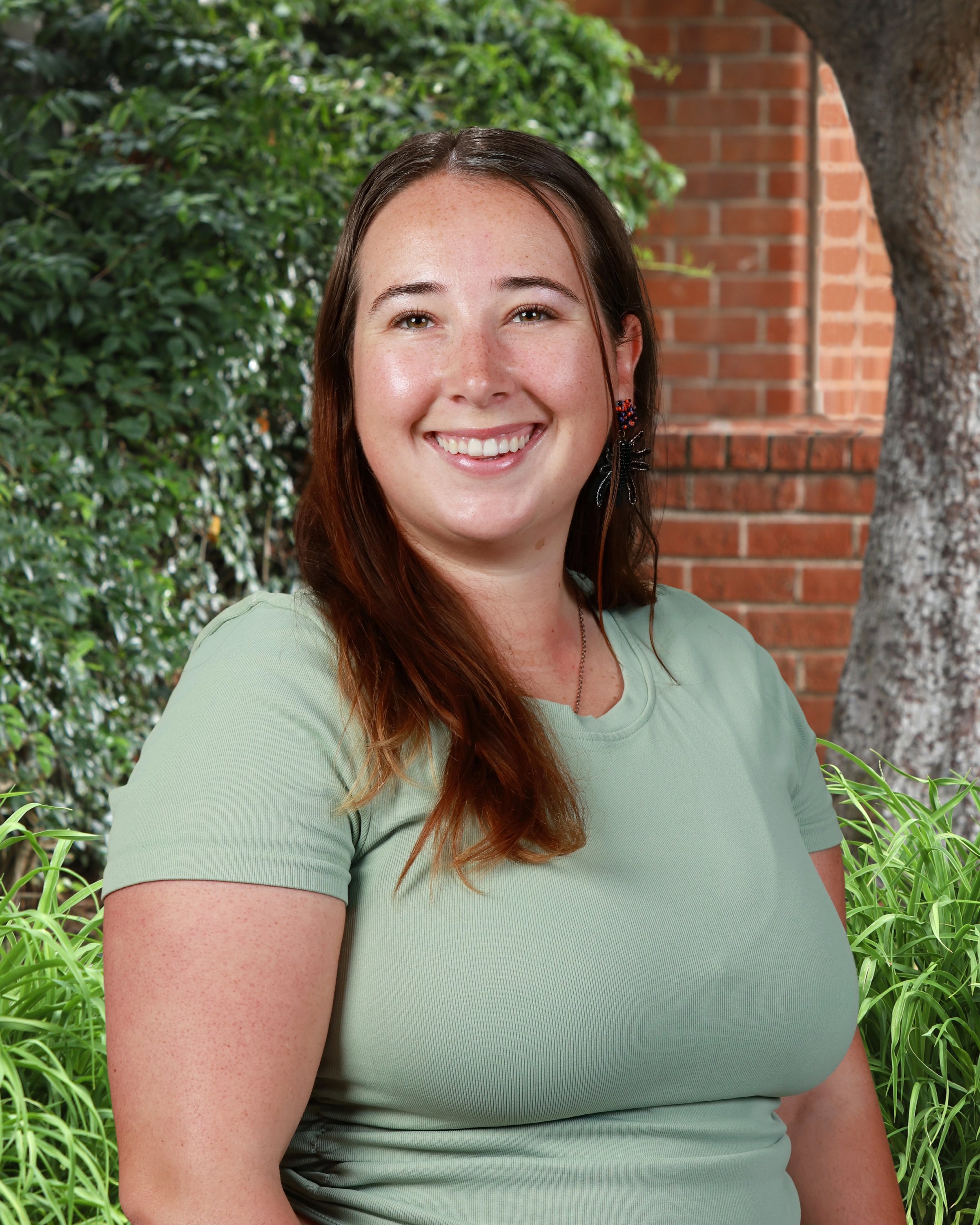 Smiling woman with brown hair and earrings outdoors with greenery and brick wall in background.