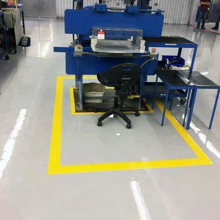 Factory workstation with a blue machine and tables inside a marked yellow safety zone on a coated industrial floor.