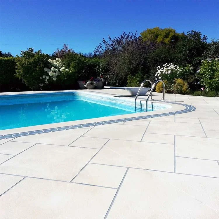 Clear blue swimming pool with a ladder surrounded by a white tiled-look coated deck and flowering shrubs.