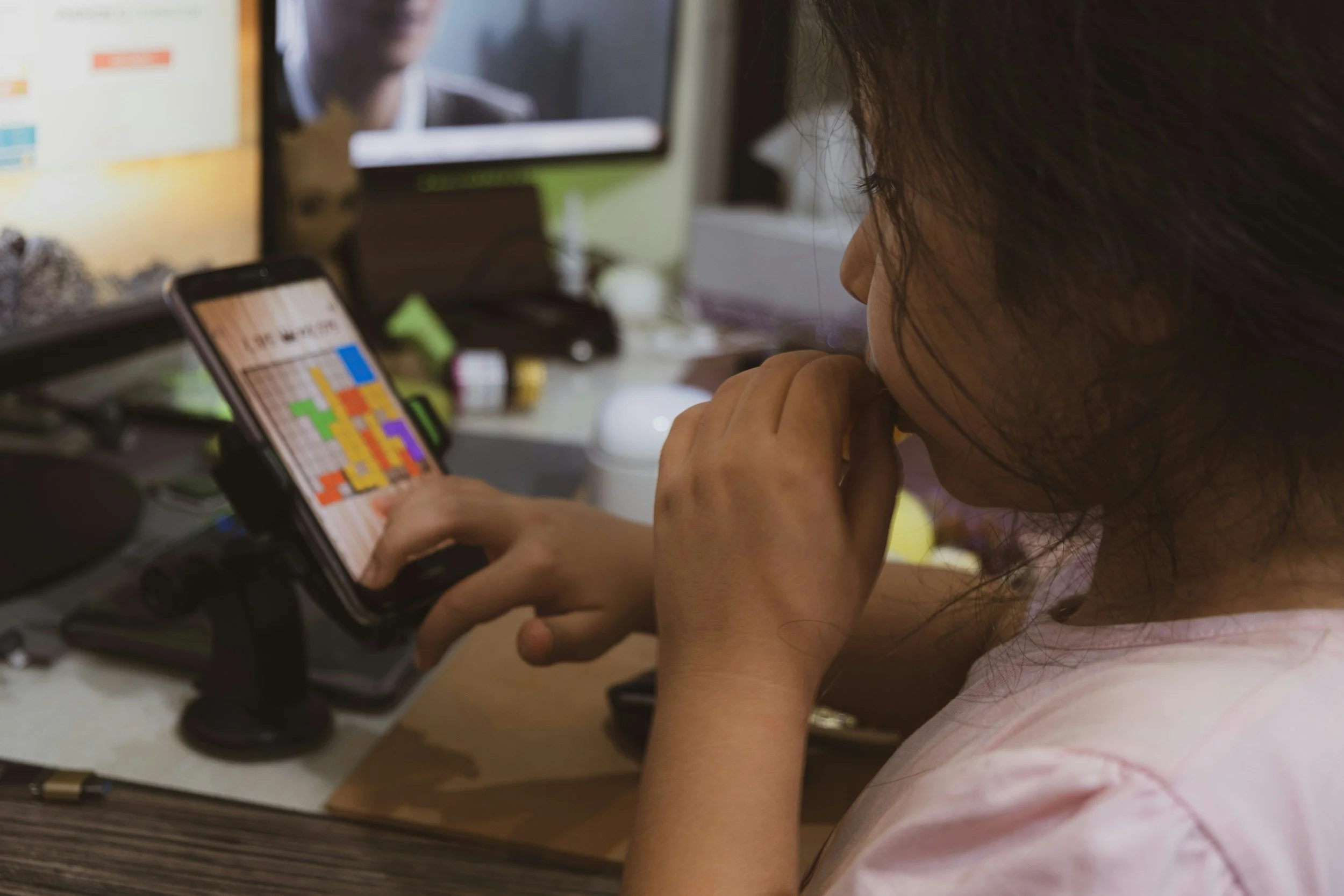 Young child focused on a smartphone game while seated at a computer desk, representing anxiety, screen dependence, and digital coping behaviors in children.