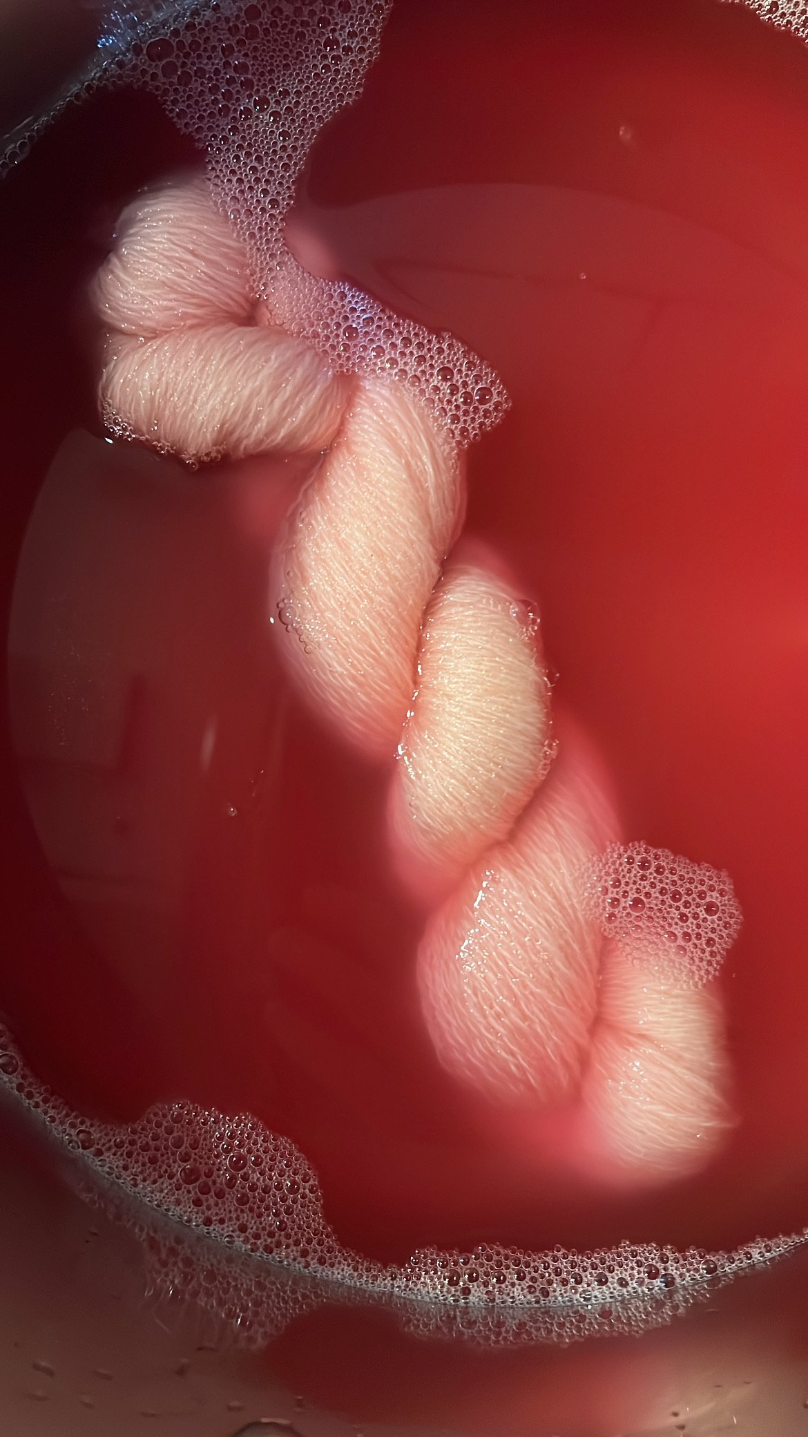 Close-up of yarn submerged in pinkish-red liquid, with bubbles on the surface.