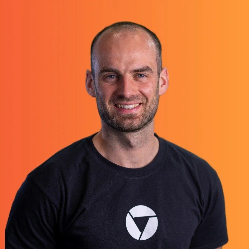 A smiling man with a beard and short hair wearing a black t-shirt with a logo, standing against an orange background.
