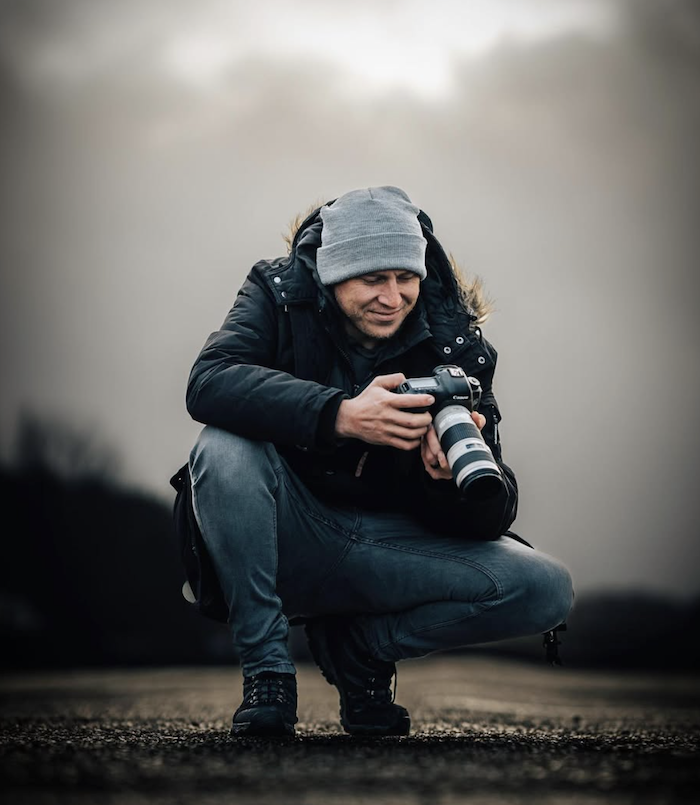 A man crouching outdoors on a dirt path, wearing a gray beanie, black jacket, and jeans, holding a professional camera with a large lens, looking at the camera's screen.