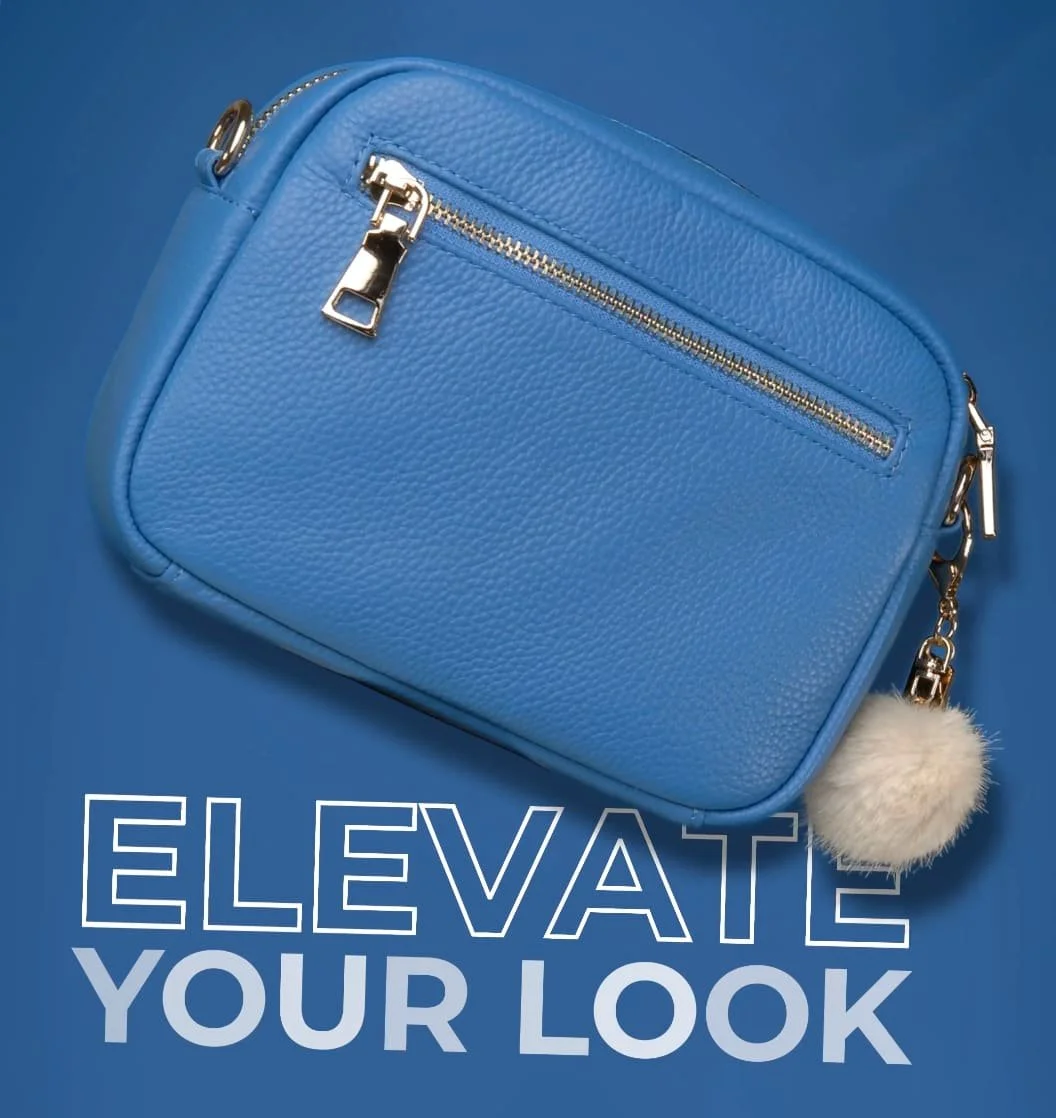 Close-up of a blue handbag with silver zippers and details, resting on a matching blue background with the words "ELEVATE YOUR LOOK" in bold white and gray text. A plush white pom-pom is attached to the handbag.