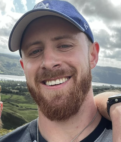 Close-up of a smiling man with a beard wearing a blue cap, outdoors with a scenic landscape of hills and water in the background.