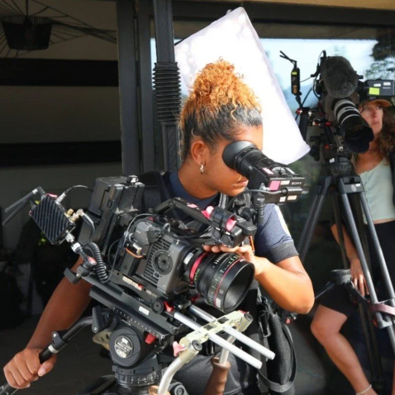 A woman operating a professional camera on a drone stabilizer, with a filmmaker and camera crew in the background.