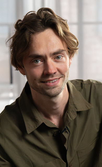 Smiling man with wavy brown hair wearing a green shirt in a bright room.