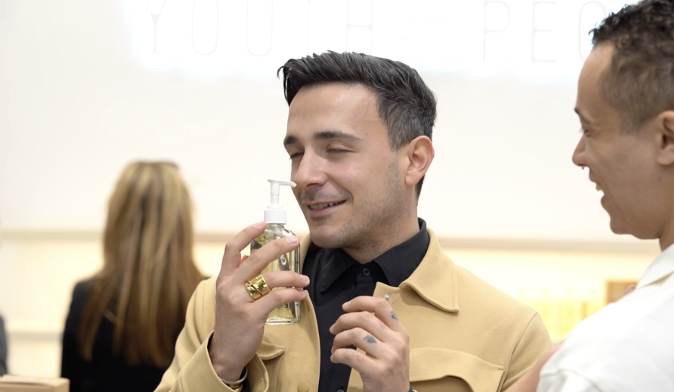 A man with dark hair, holding a pump bottle of sanitizer close to his face with a smile, talking with another person in a white coat in an indoor setting.