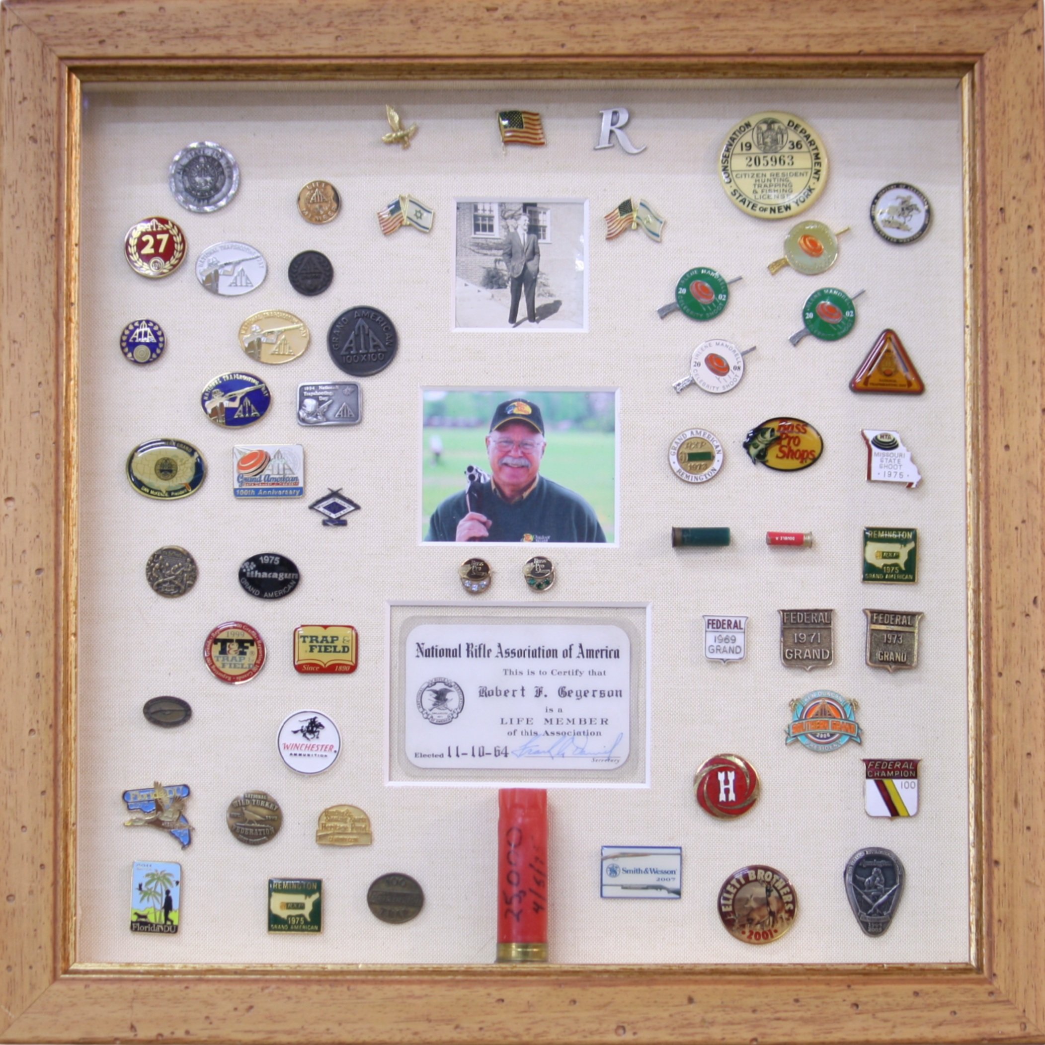 A shadow box displaying various lapel pins, two photographs, a certificate, and a shotgun shell centered at the bottom.