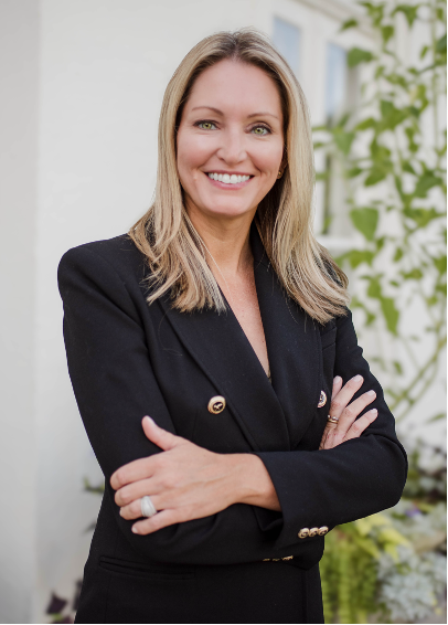 A smiling woman with blonde wavy hair wearing a plaid shirt, leaning against a stone railing outdoors.