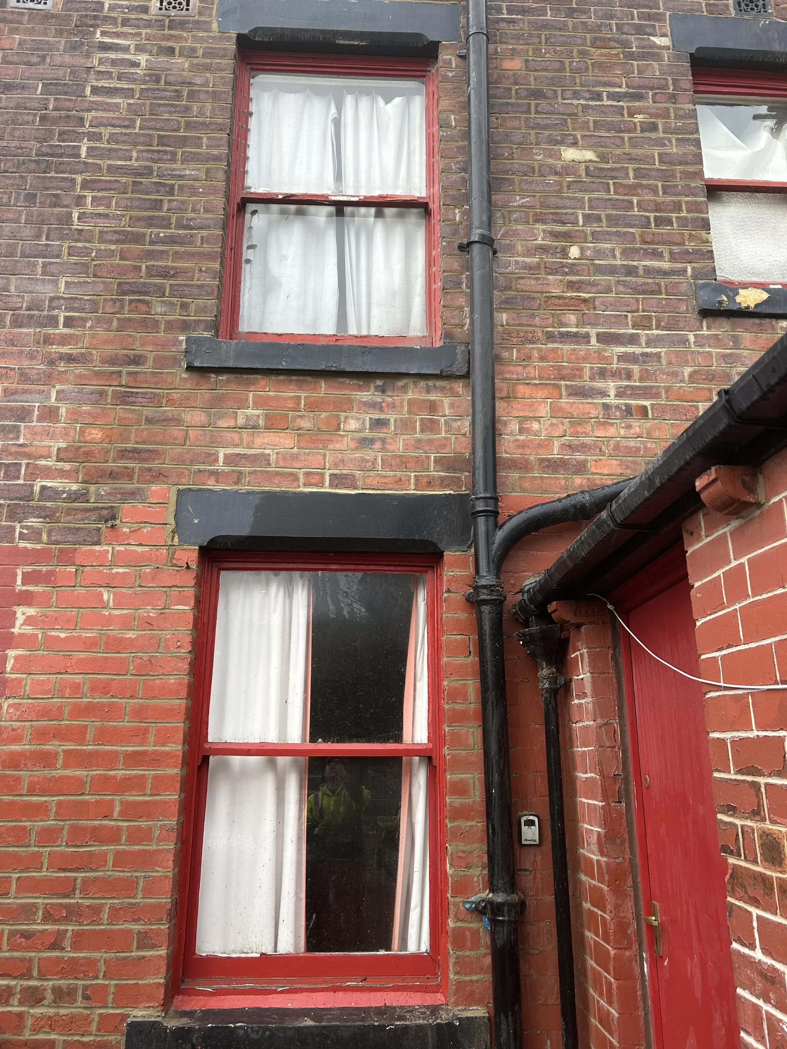 Two brick building windows with white curtains and red frames, black exterior trim, black downspout and pipe, and red door with a brass handle.