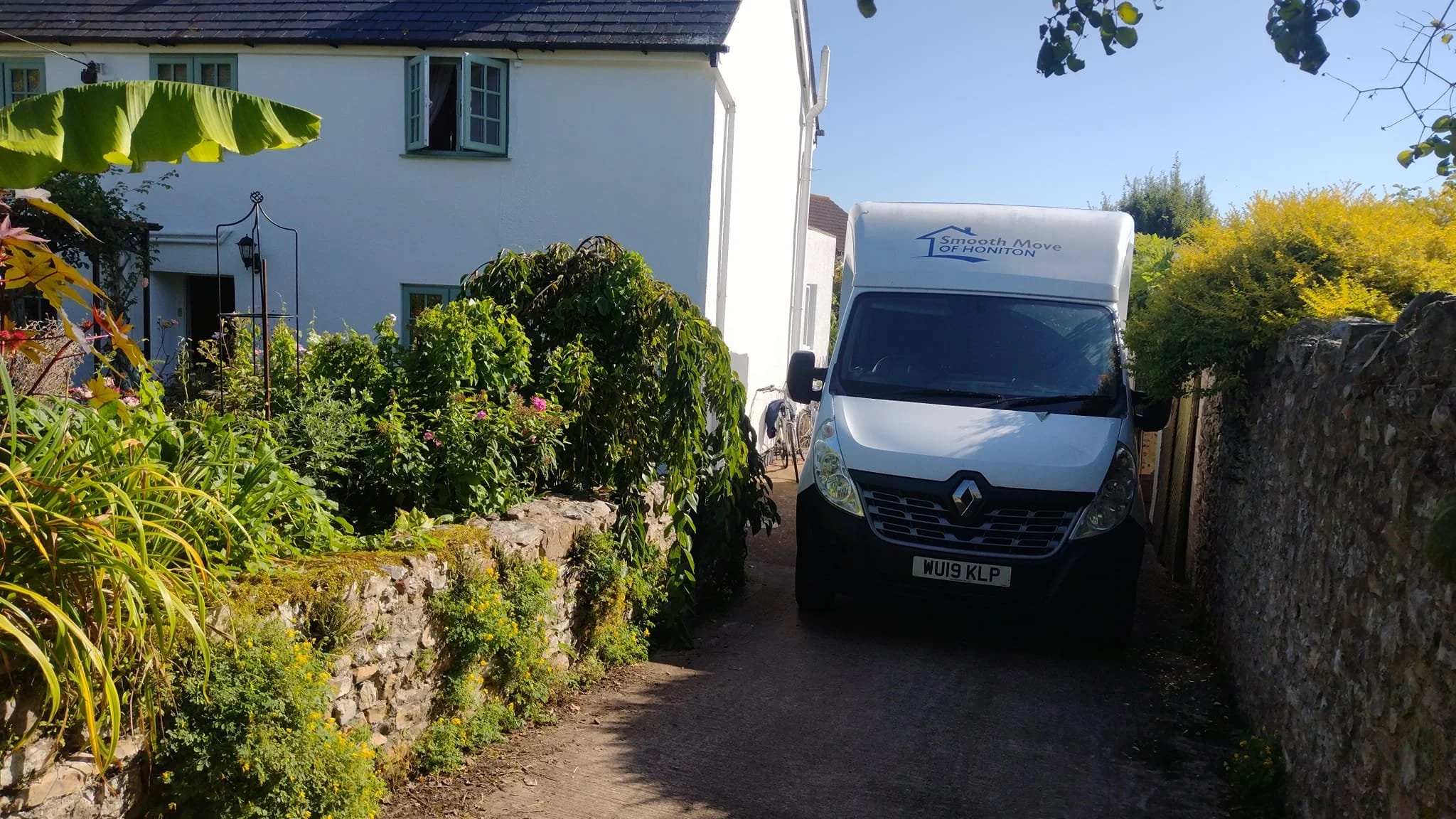 A white delivery van parked in a narrow alleyway between a garden and a stone wall on a sunny day.