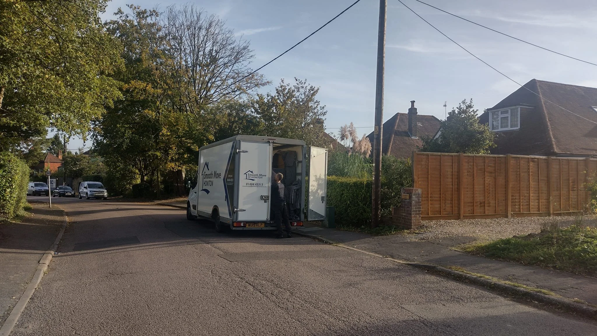 a man loading boxes into a white moving van on a suburban street with houses, trees, and parked cars nearby