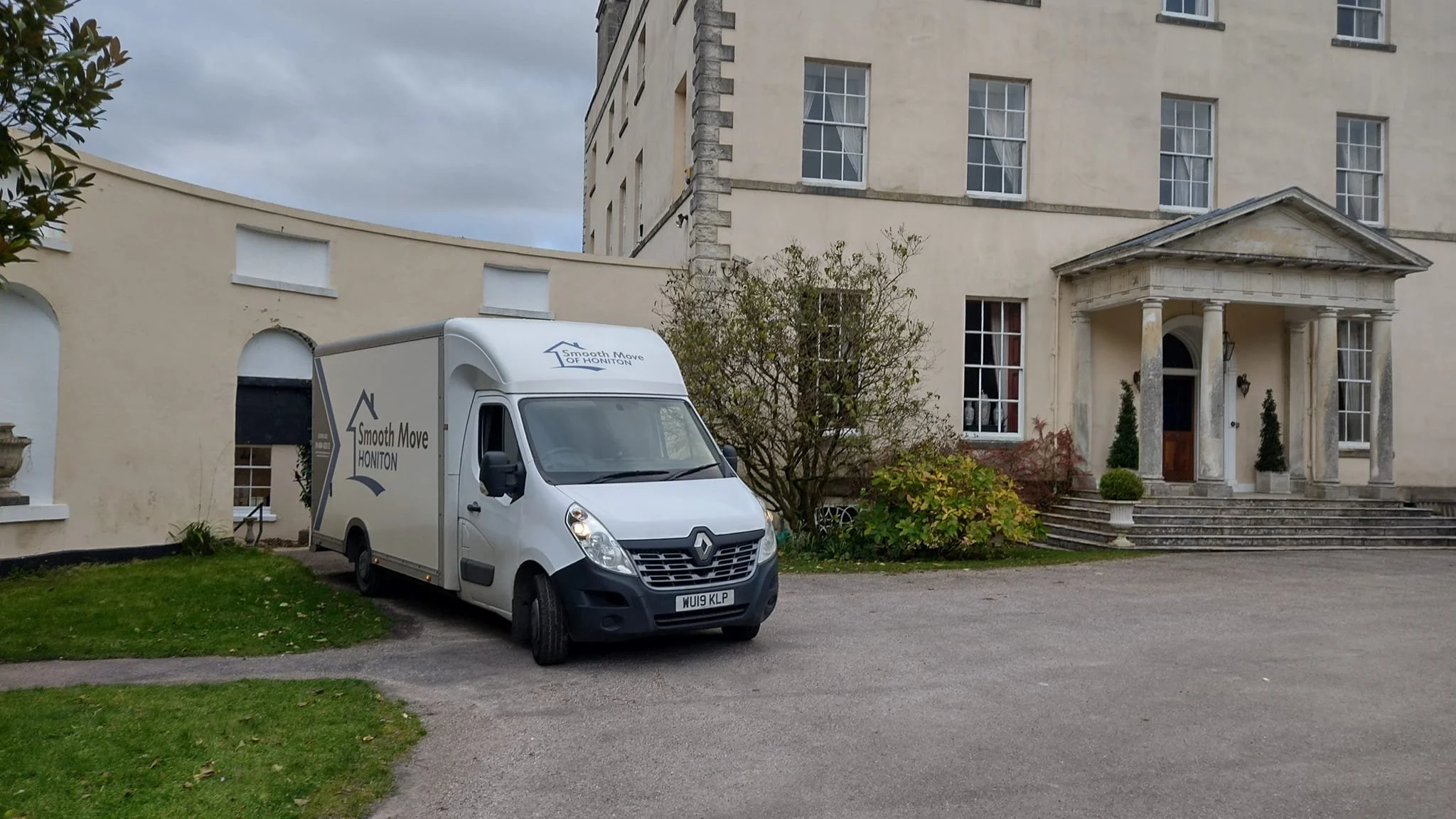 A white moving van with logo 'Smooth Move HONITON' parked on a driveway in front of a large beige historic building with a portico entrance, steps, and multiple windows, with trees and shrubs nearby.