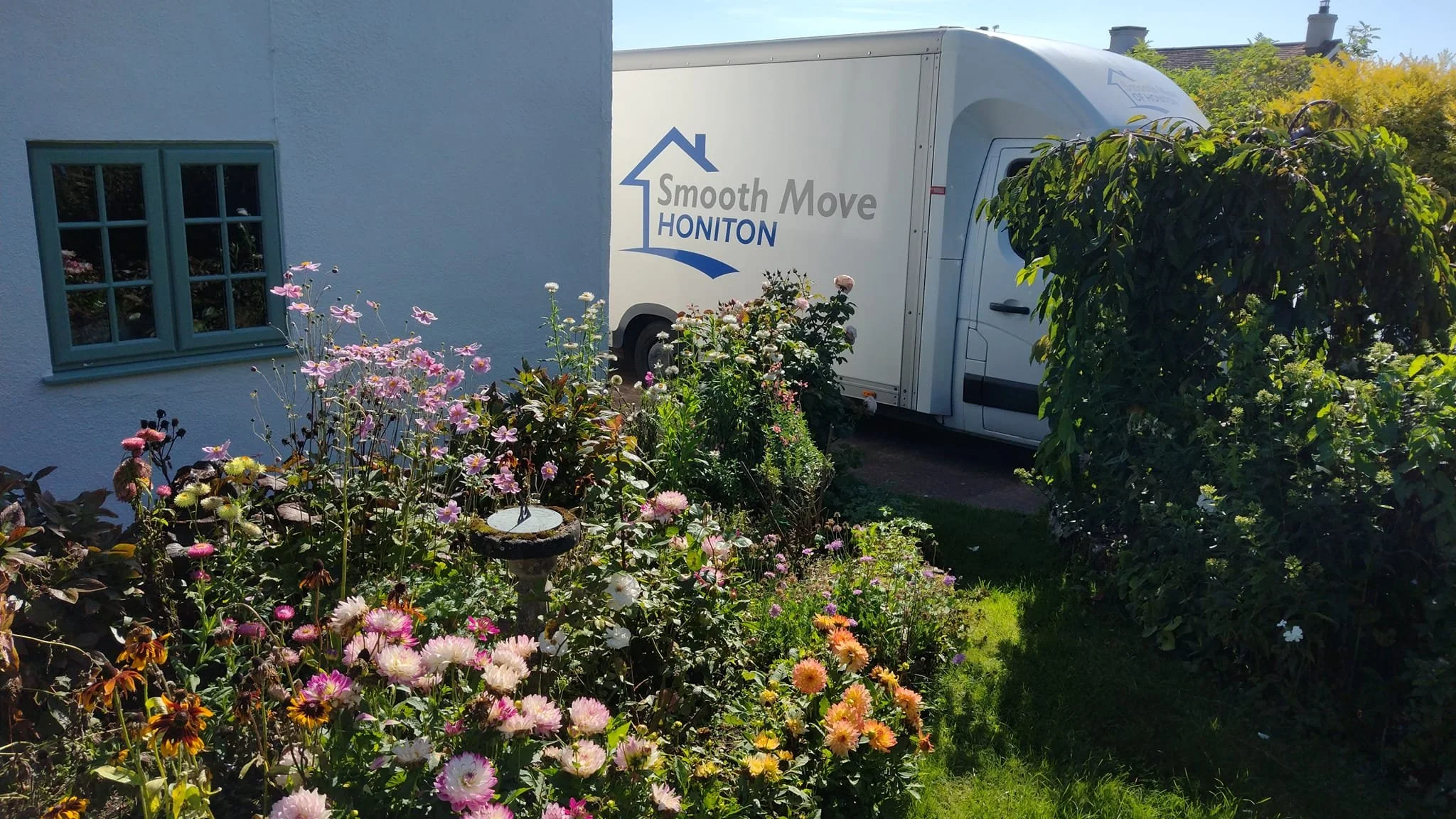 A white moving company truck parked near a house with green window frames is partially obscured by lush garden with pink, white, and yellow flowers and greenery.