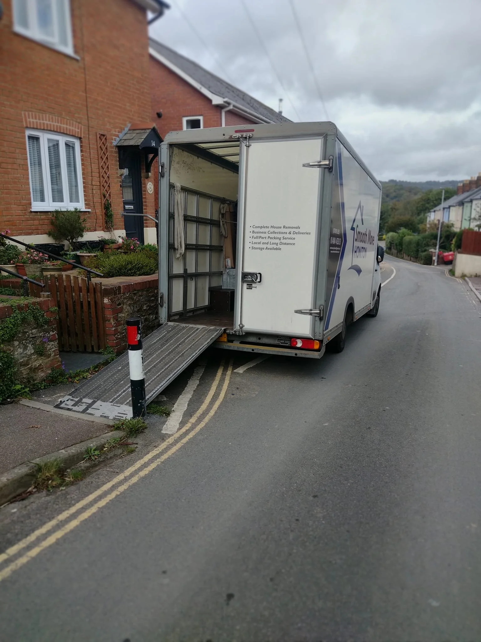 A moving truck with side door open and a ramp extended onto a residential street with houses and cloudy sky.