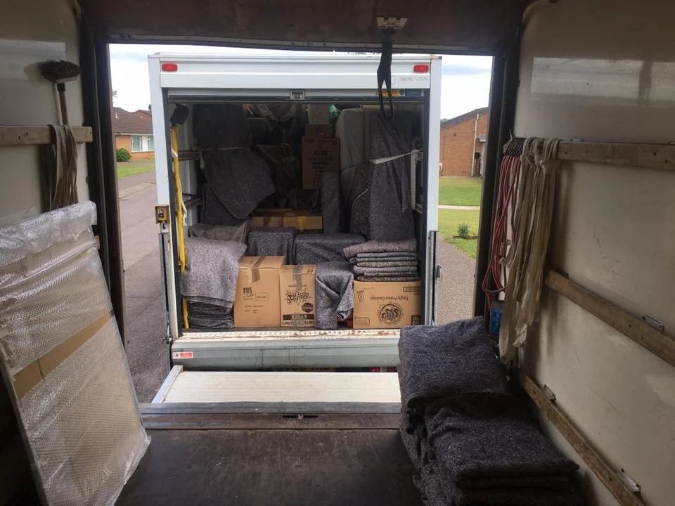 The interior of a moving truck filled with boxes and furniture, with a view of a suburban neighborhood outside.