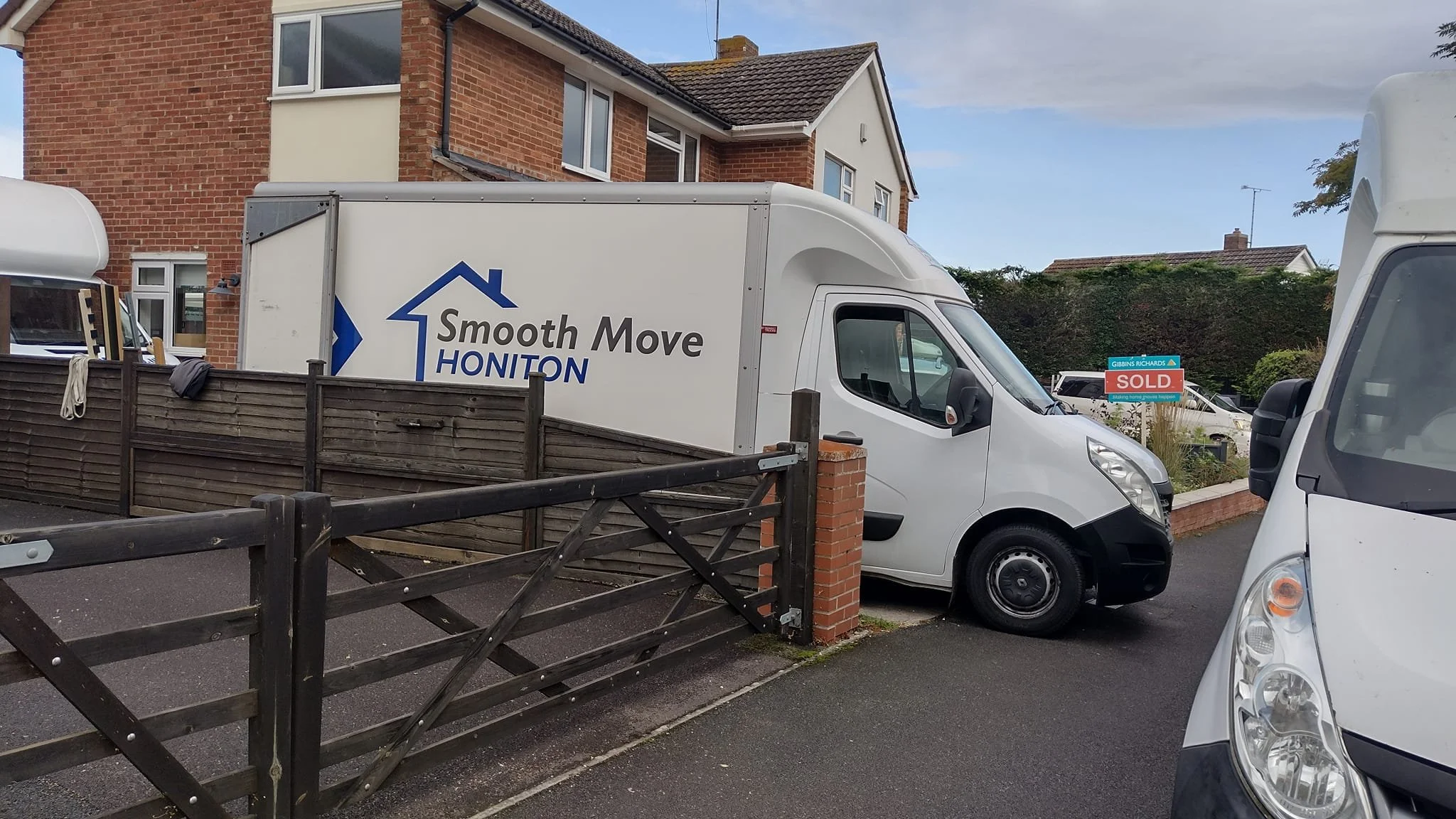 White moving truck with 'Smooth Move Honiton' logo parked in front of a house, with a 'Sold' sign in the yard and a black wooden fence.