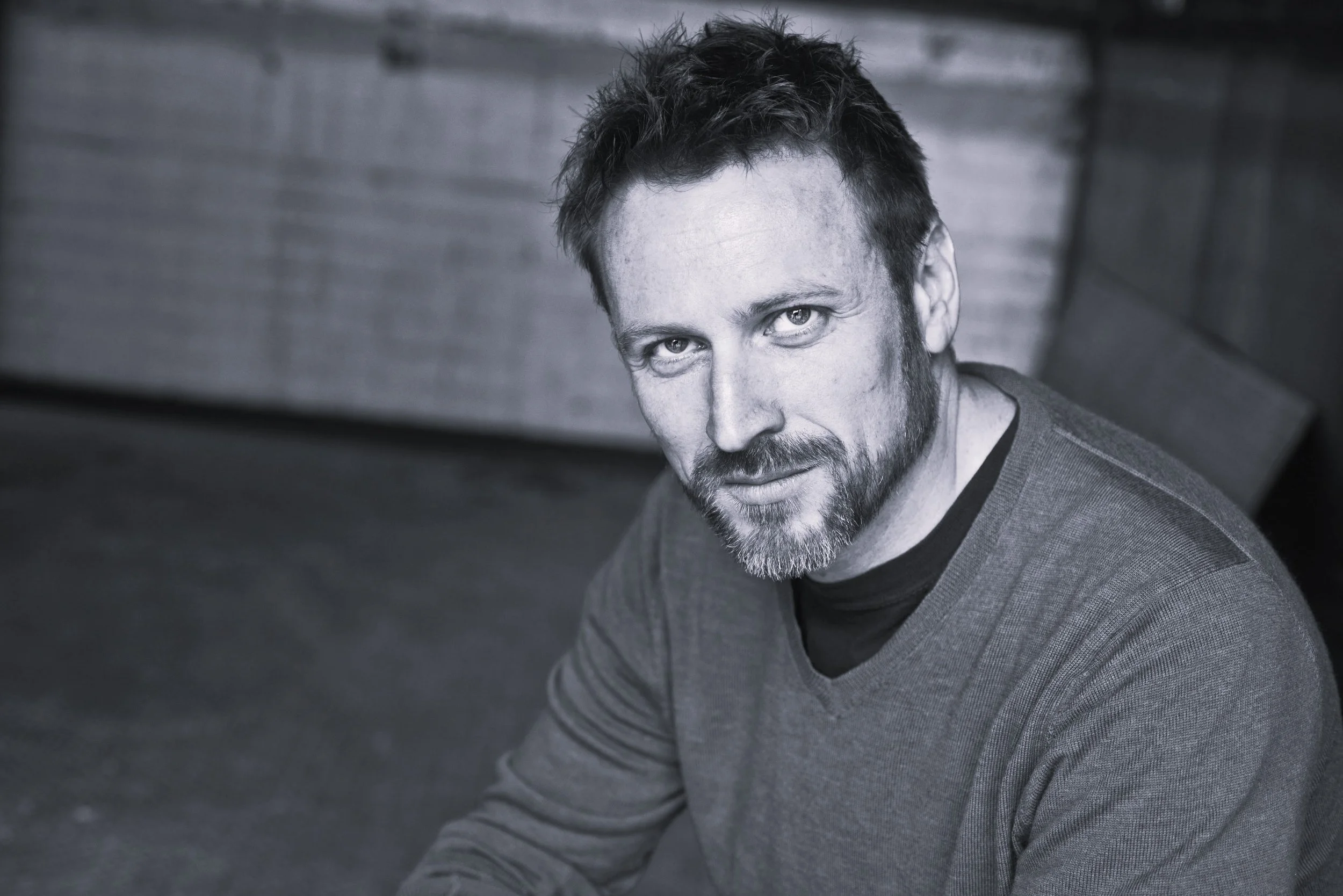 A black-and-white portrait of a man with short dark hair, a beard, and mustache, wearing a sweater, sitting outdoors near a wooden structure, looking into the camera.