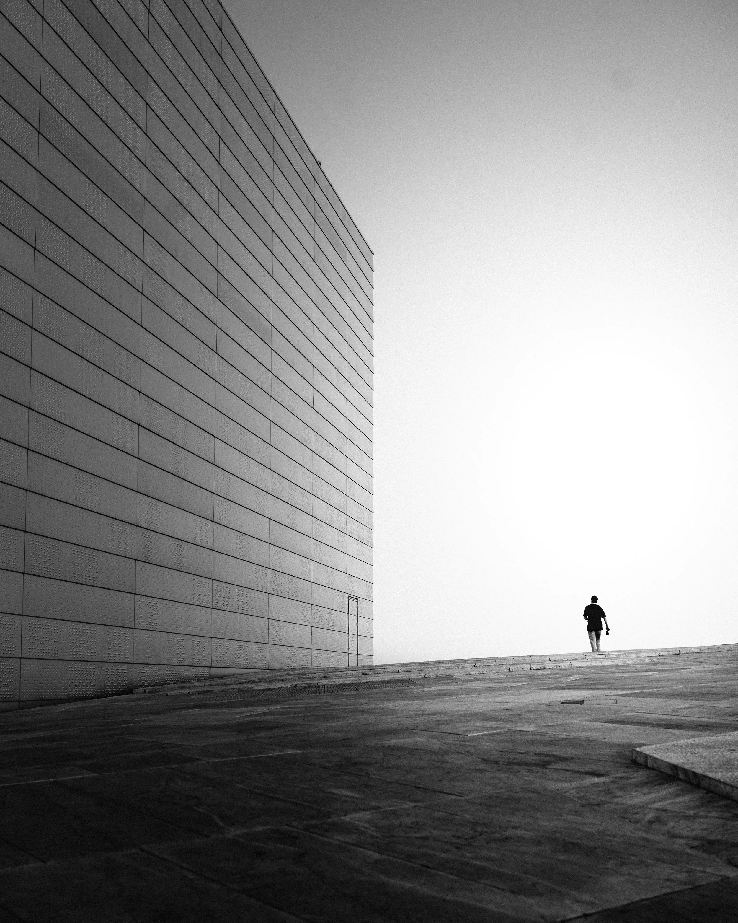 A black and white photo of a large modern building on the left, with a person walking away in the distance on an open paved area.