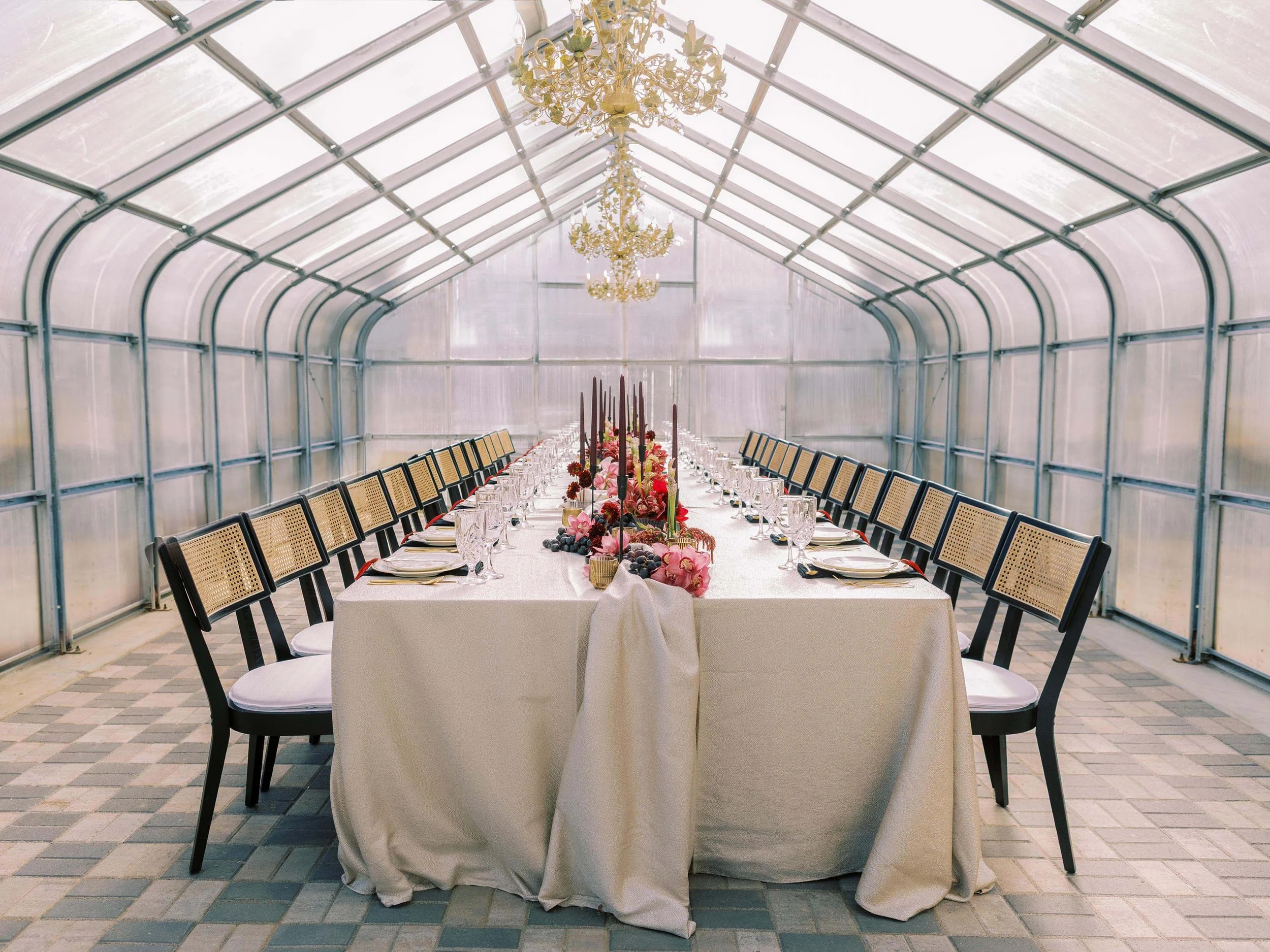long party table in a greenhouse with sleek rattan chairs, tall burgundy candlesticks, and a fruit and floral table runner