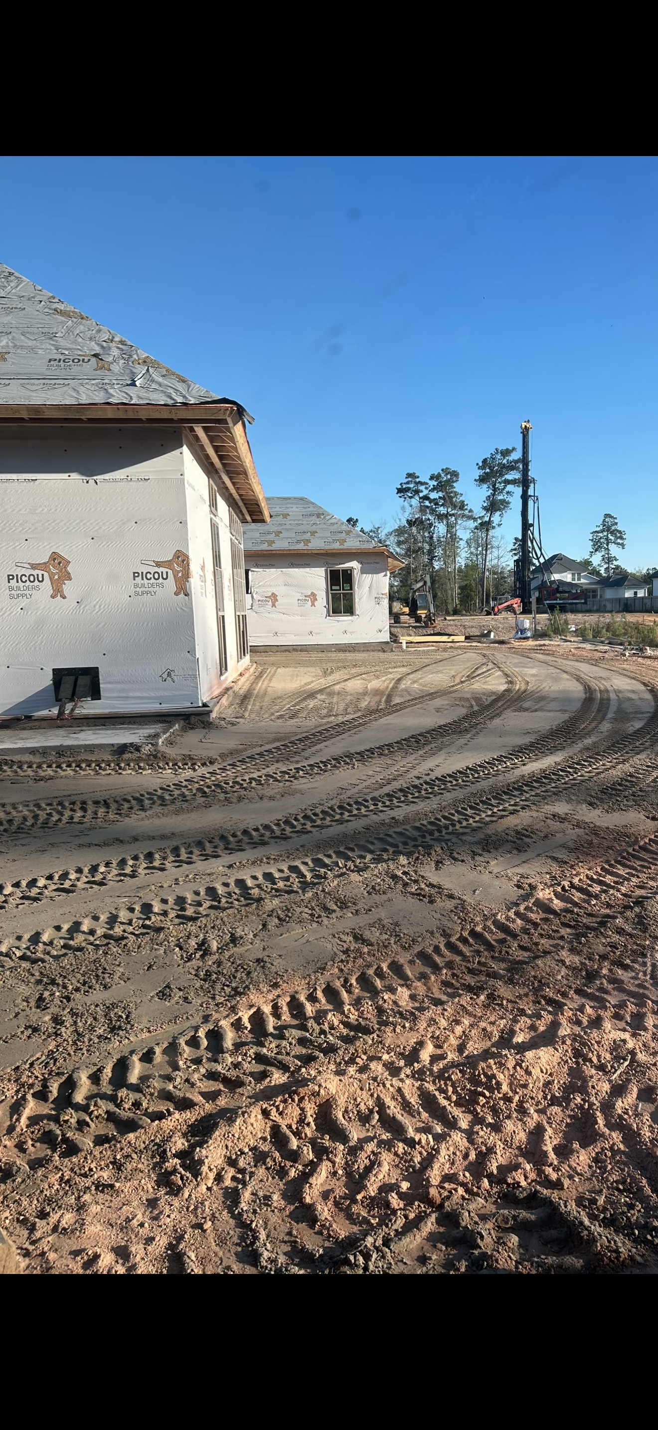 A dirt road extending into a wooded area with a pile of logs on the left and a red Kubota skid-steer parked partially visible on the right.