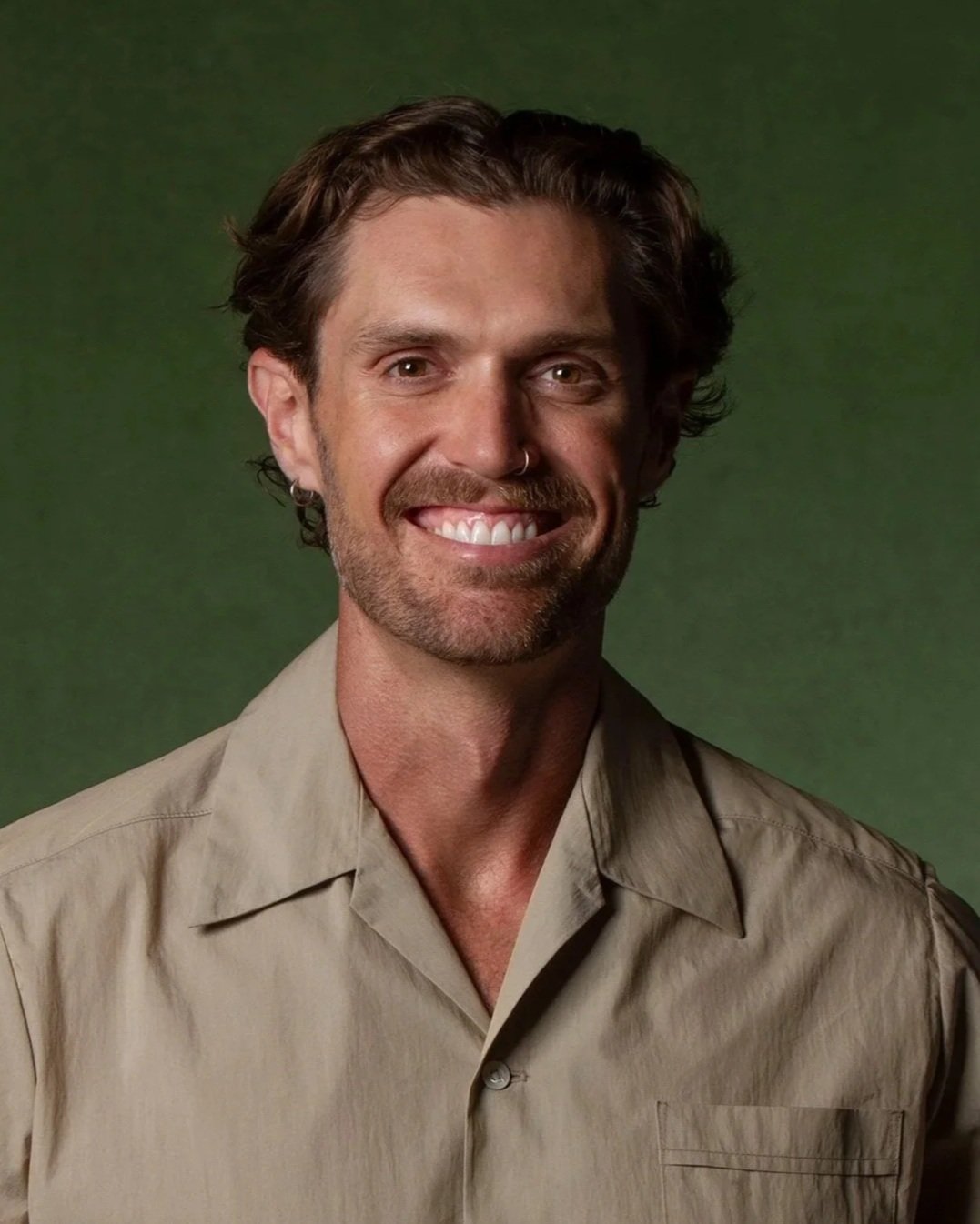 A smiling man with wavy brown hair, a beard, and a nose ring, wearing a beige button-up shirt against a green background.