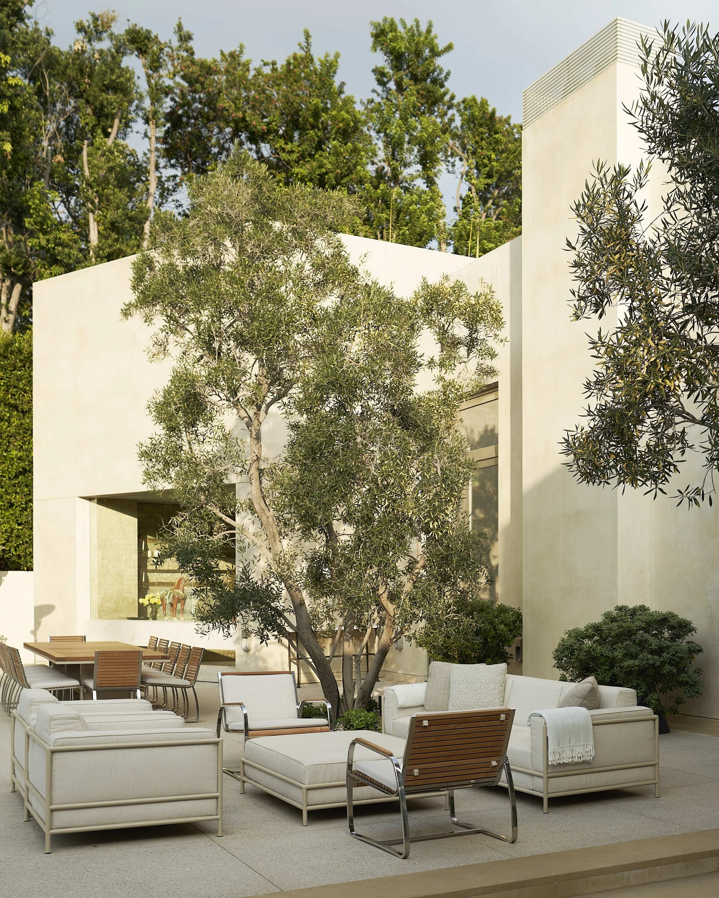 Modern outdoor patio with white cushioned furniture, trees, and a beige building surrounded by greenery.