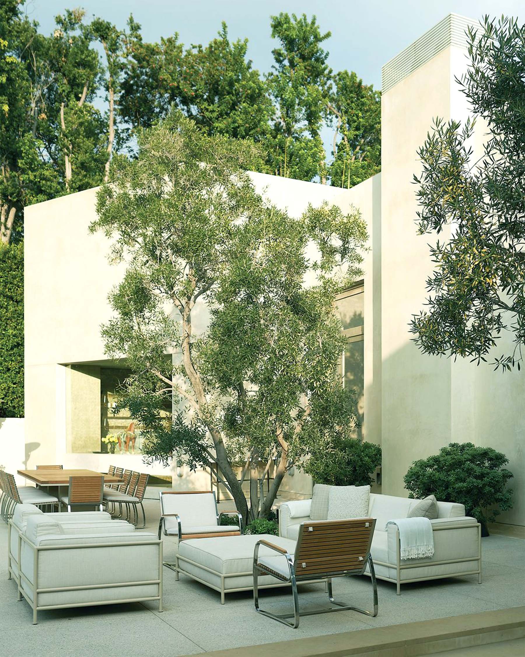 Modern outdoor patio with white furniture, a large tree, and lush greenery surrounding a sleek white house.
