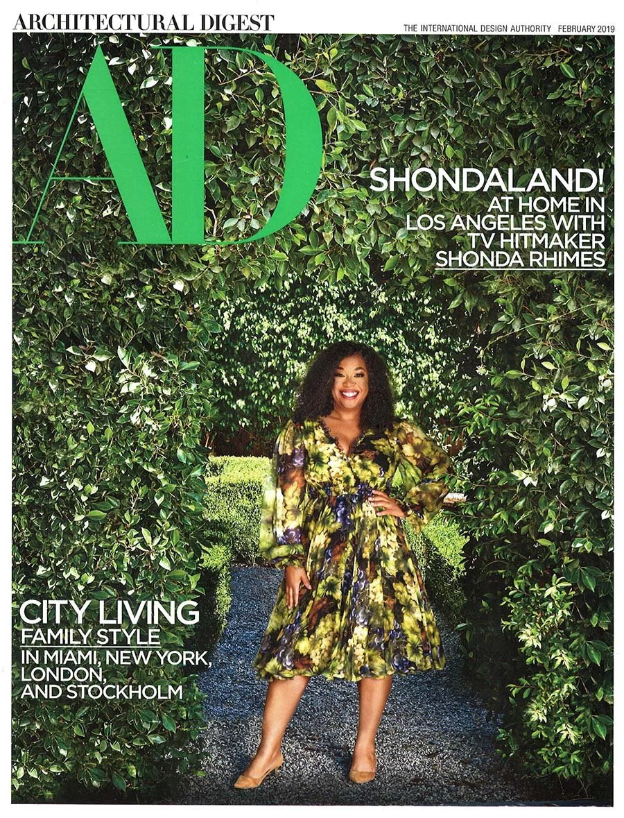 A woman standing in a lush, green garden archway made of dense foliage. She is wearing a colorful, floral-patterned dress and beige heels, smiling with one hand on her hip.