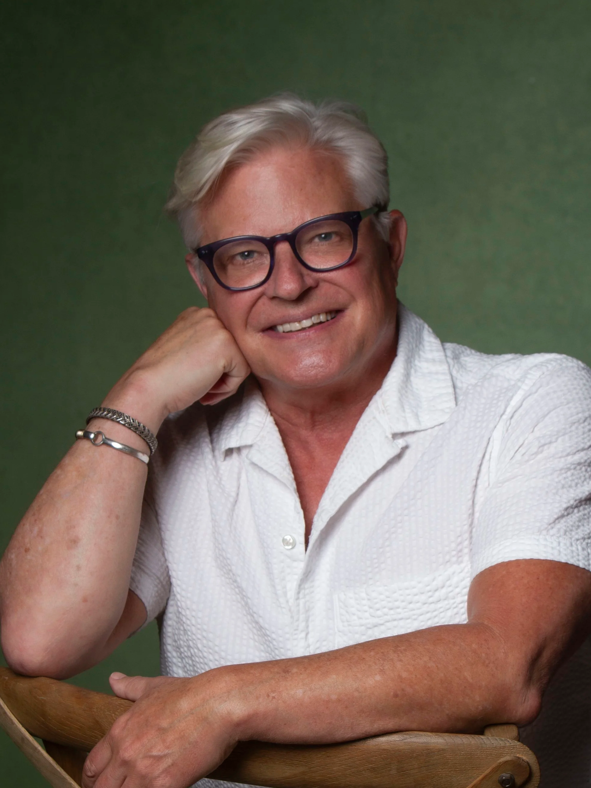 A smiling elderly man with white hair and glasses, wearing a white shirt, resting his chin on his hand against a green background.