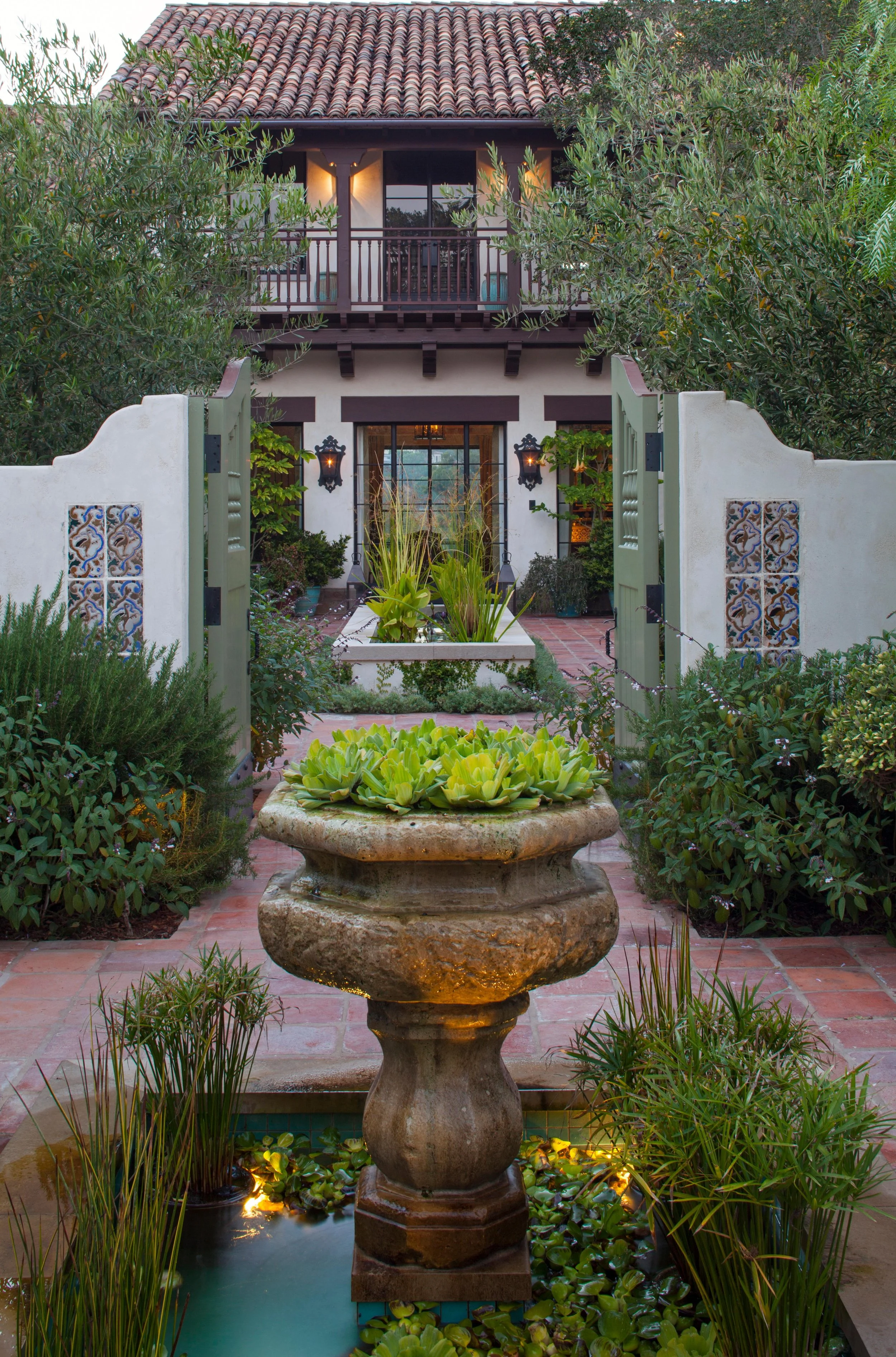A courtyard garden with a stone fountain surrounded by lush plants and a red-tiled house with a balcony and outdoor living space.