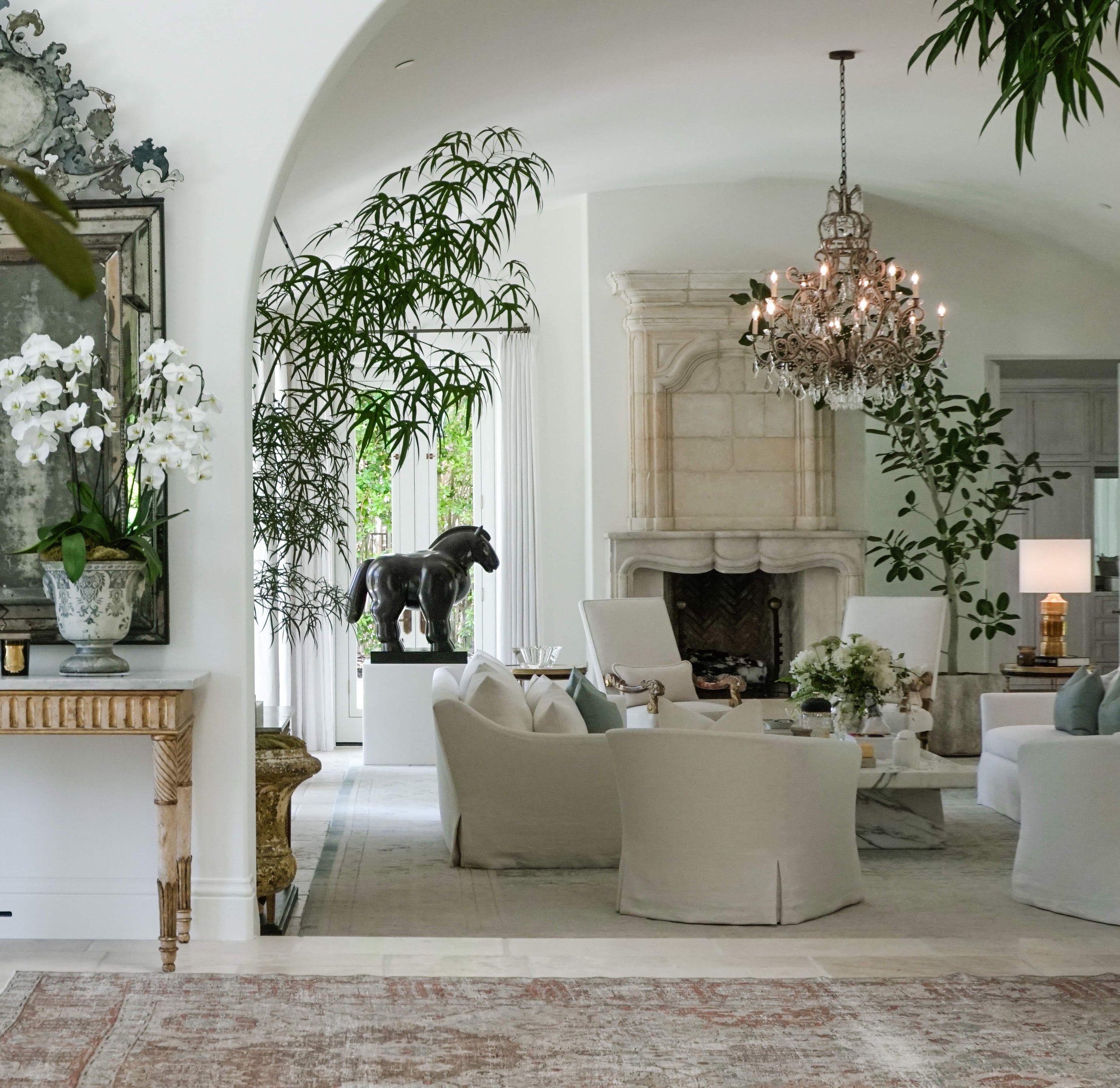 Elegant living room with white furniture, a fireplace, chandelier, potted plants, decorative horse statue, and large windows with curtains.