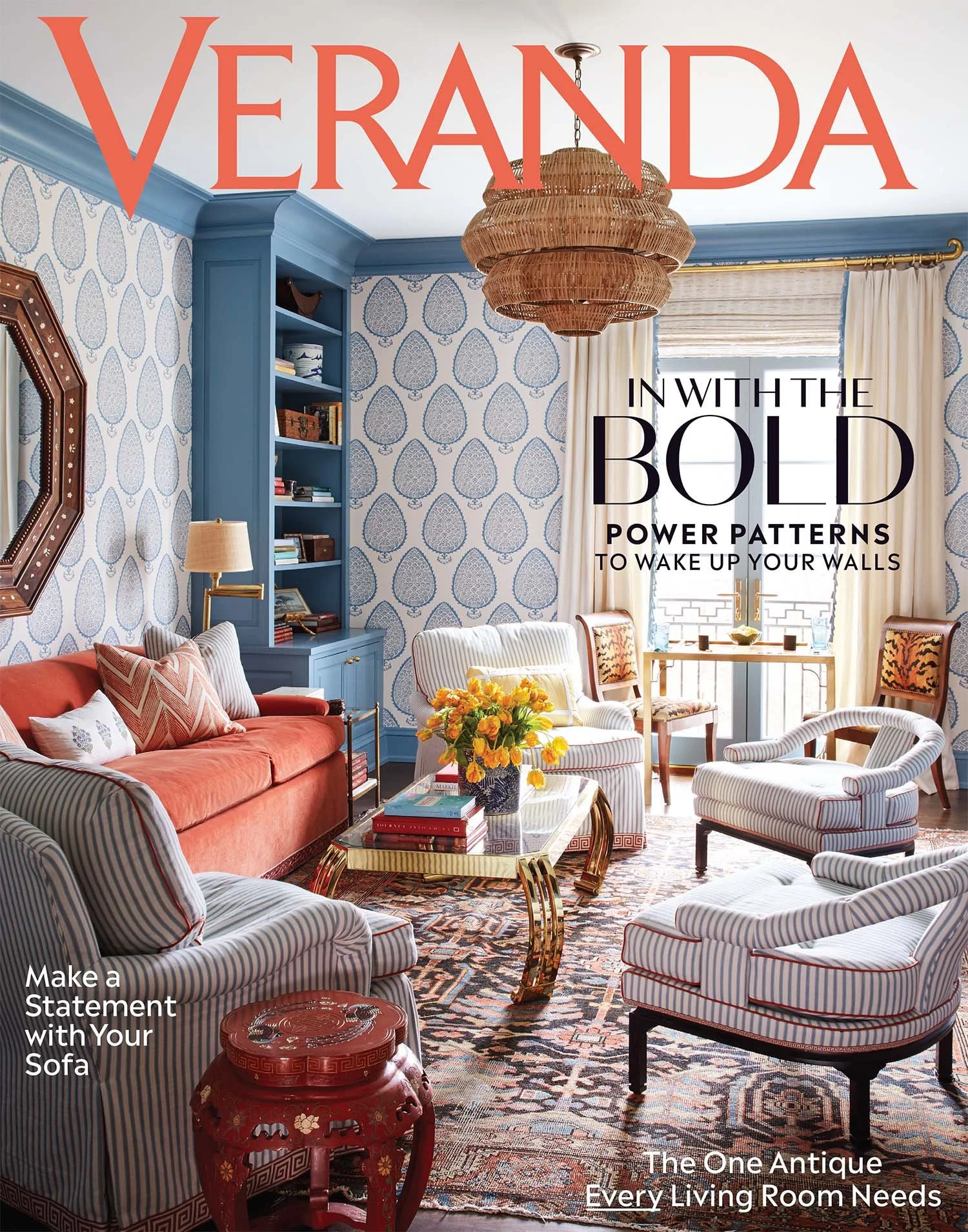 Living room with blue and white wallpaper, orange sofa, striped armchairs, glass coffee table with books and a flower vase, patterned carpet, bookshelf, window with curtains, and a woven hanging light fixture.