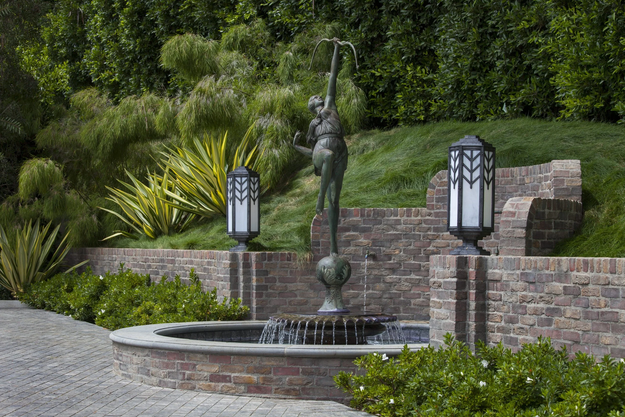 Outdoor garden fountain with a bronze statue of a woman holding a bow, surrounded by greenery, brickwork, and lamps.