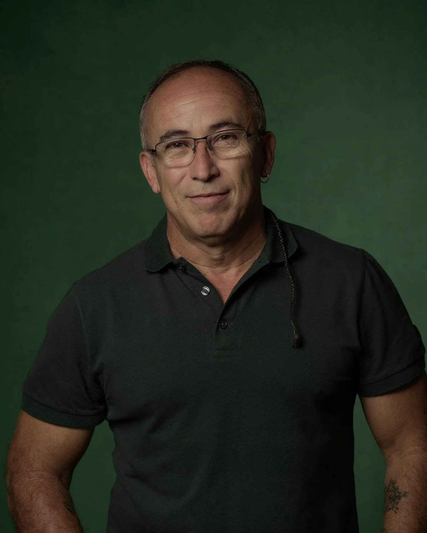 Portrait of a middle-aged man wearing glasses and a black polo shirt, standing in front of a green background.
