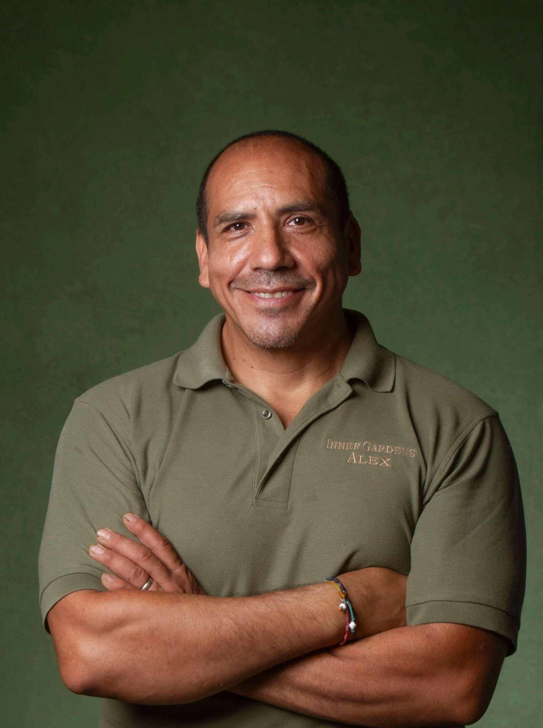 A man with a friendly smile, short dark hair, and a goatee, wearing an olive green polo shirt with the words 'Inner Gardens Alex' embroidered on it, standing with arms crossed against a green background.