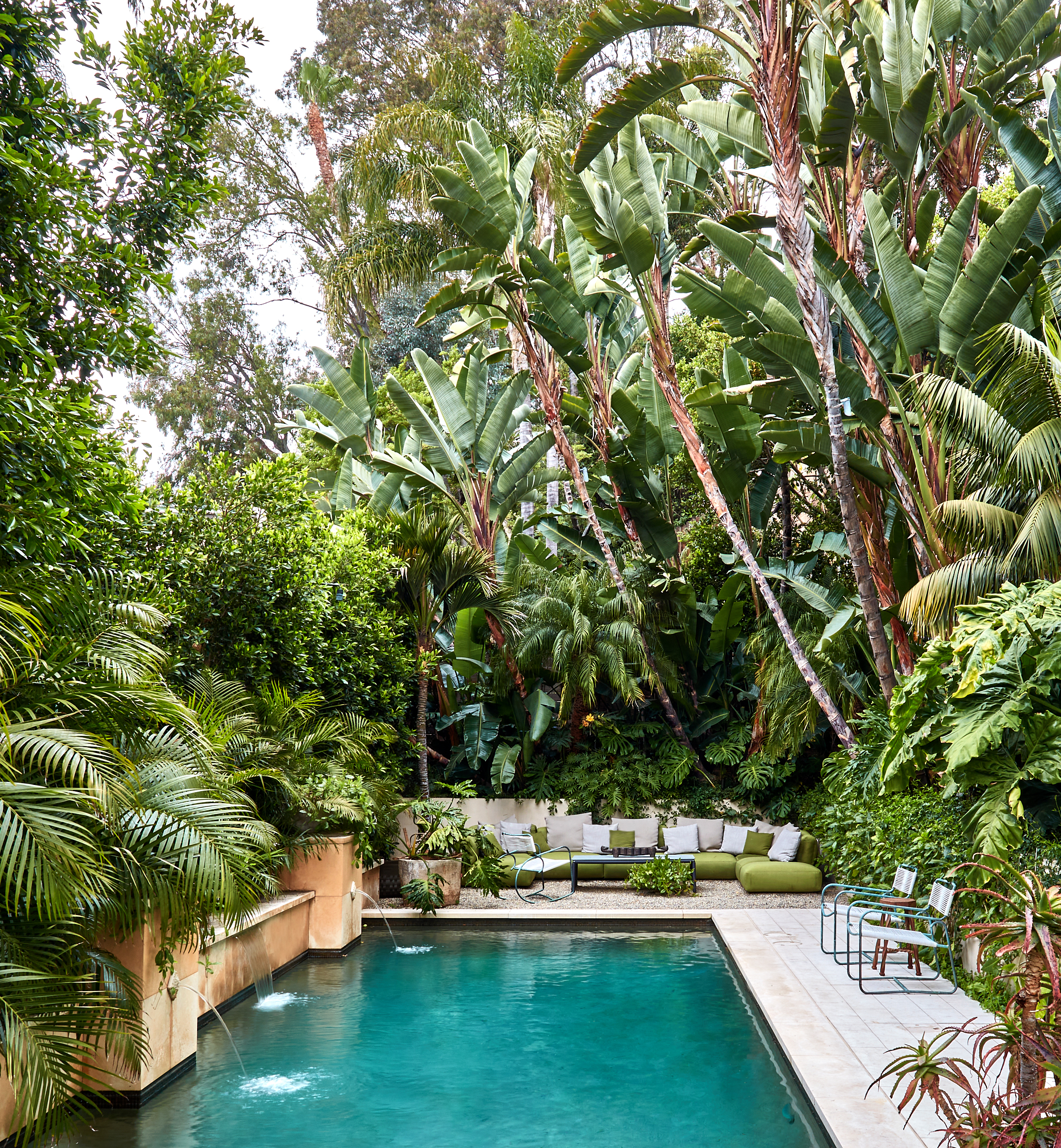 A backyard swimming pool surrounded by lush tropical plants, with a long outdoor sofa with white cushions and pillows at the back, a small black coffee table, and a few chairs and tables on the deck.