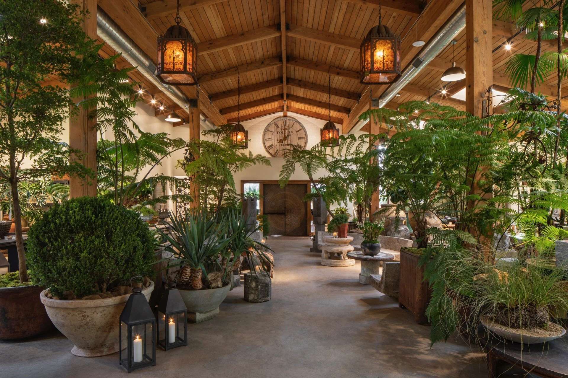 Interior of an indoor garden space with green plants in pots, lanterns with candles on the floor, wooden ceiling with hanging lights, and a large wall clock above a door.