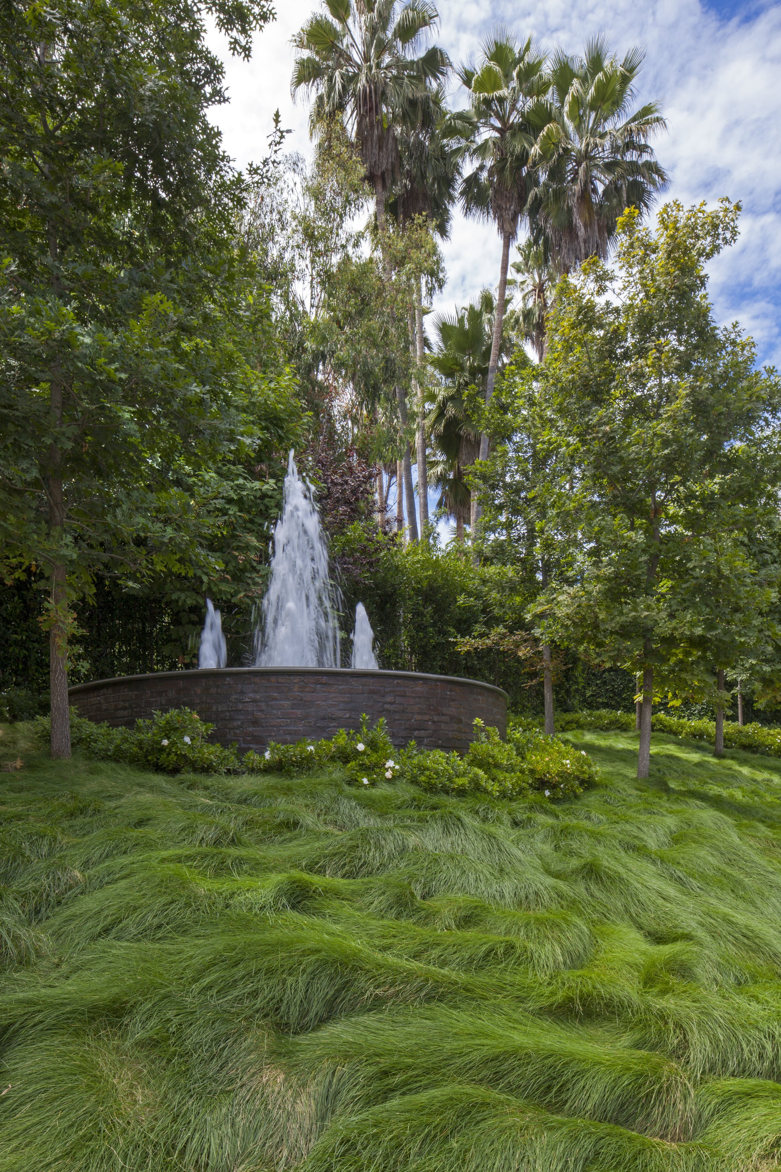 A lush garden with a fountain surrounded by green grass, trees, and bushes, under a partly cloudy sky.